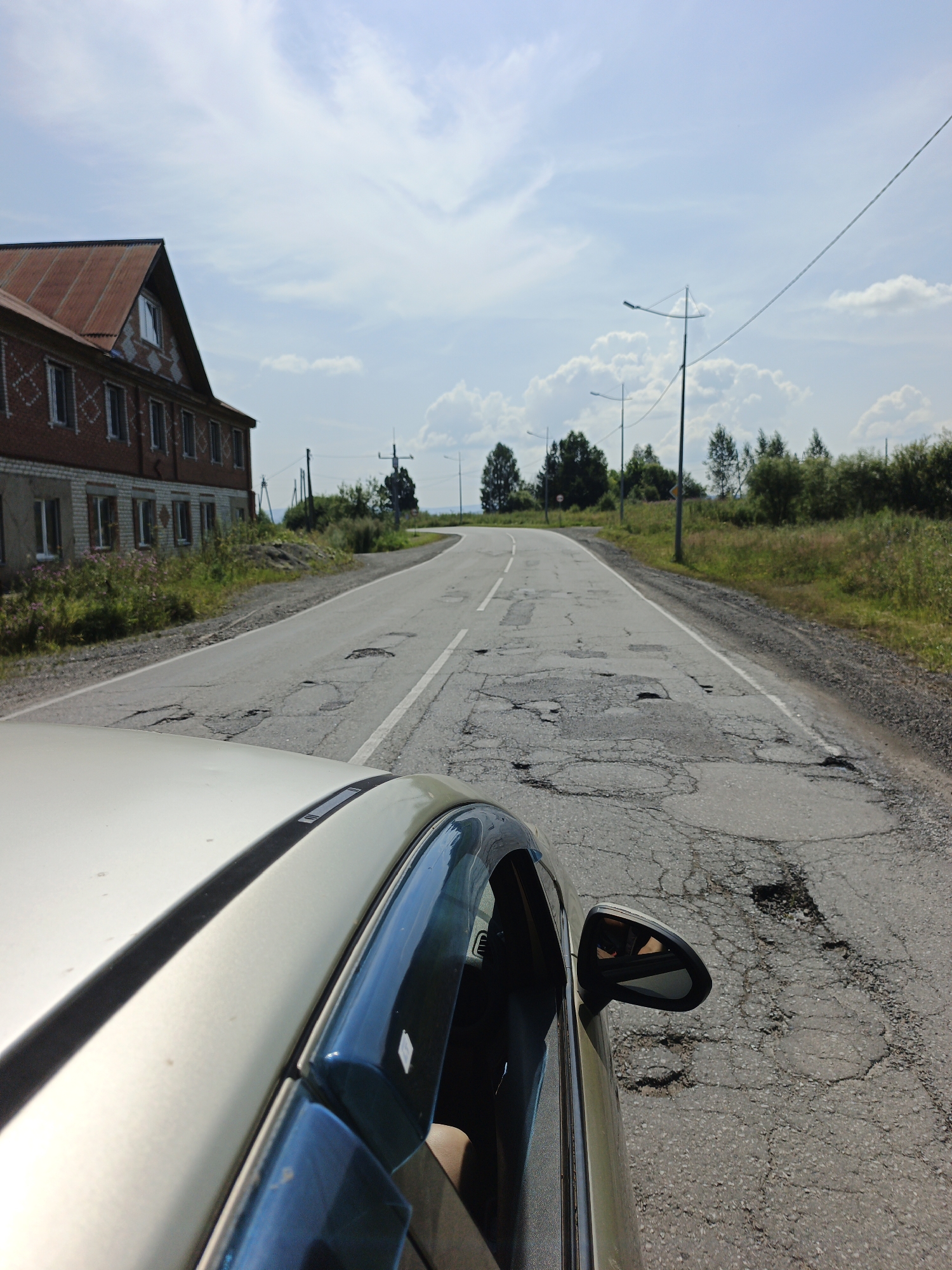 The image shows a view from the passenger side of a car, looking forward along a rural road in Medvedyovka, Russia. The road is paved but heavily damaged with numerous potholes and cracks. On the left side of the road, there is a two-story brick building with a red, corrugated metal roof. The building has several windows and a decorative pattern on its upper facade. Adjacent to the building is a patch of green vegetation and some pink wildflowers. The road curves to the right and continues into the distance, flanked by green trees and bushes on both sides. Power lines run parallel to the road, supported by utility poles, and several streetlights are visible. The sky is blue with scattered white clouds. The car itself is visible in the foreground, with its light-colored hood and the reflection of the sky and trees in its side window. A side mirror is also visible, reflecting the driver's arm. The overall scene suggests a journey through a rural area with challenging road conditions.