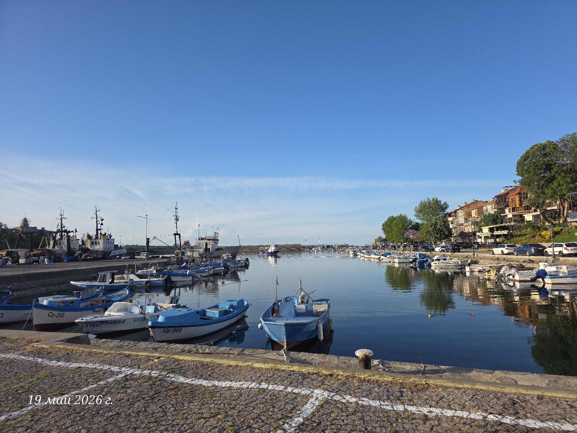 The image depicts a harbor scene in Sozopol, Bulgaria. A body of water fills the center, reflecting the clear blue sky. Numerous boats, both small fishing vessels and larger ships, are docked along the harbor. The boats are a mix of white and blue, some with Cyrillic lettering. A cobblestone pier runs along the bottom of the frame, marked with a white line. Buildings are visible on the right side of the harbor, and cars are parked along the road. The date "19 май 2026 г." is imprinted on the bottom left corner.