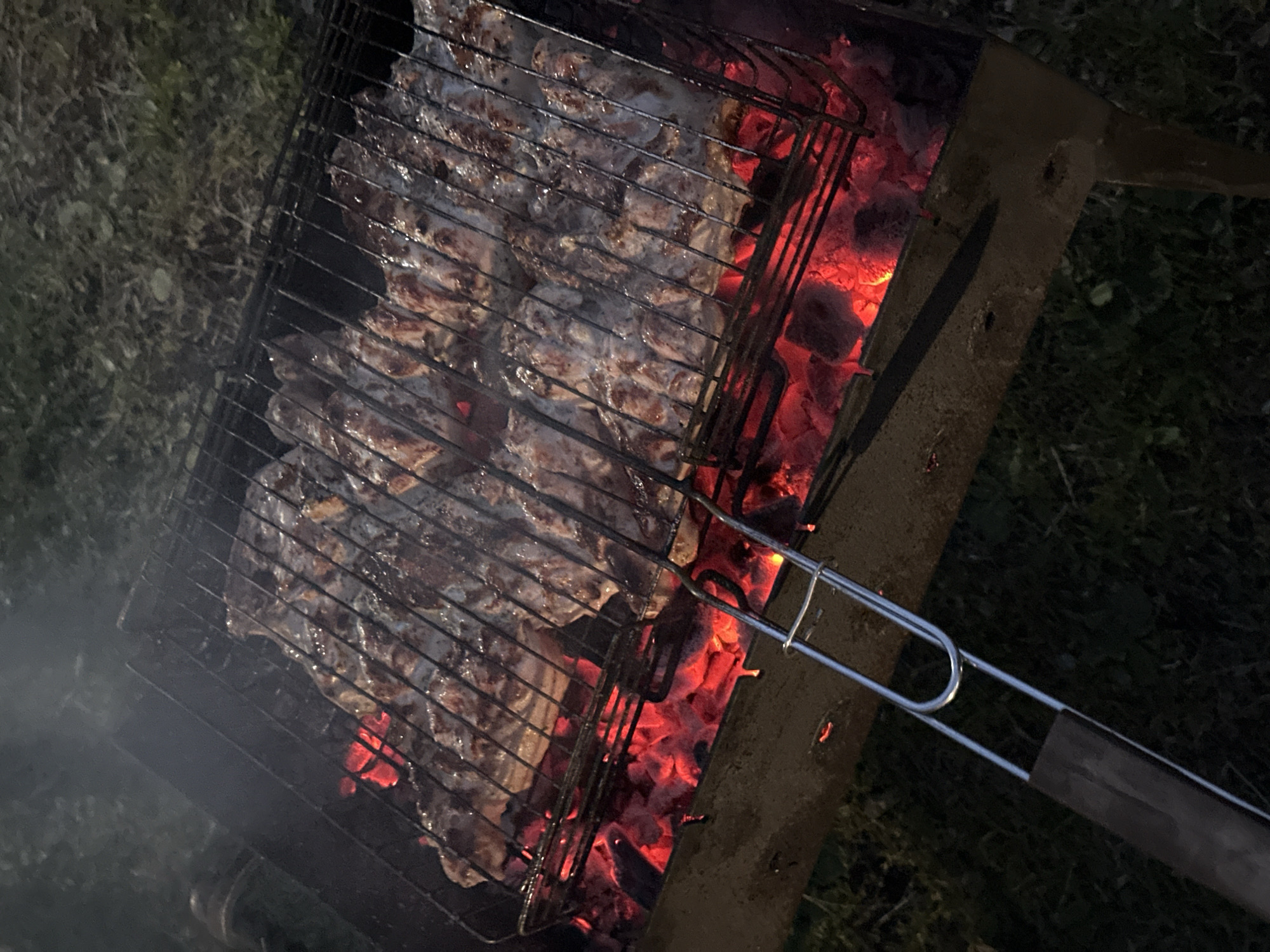 The image depicts food being grilled over hot coals in an outdoor setting. The grill, filled with pieces of meat, sits atop a metal structure containing the coals. A metal grilling tool is visible. The ground surrounding the grill is dark, possibly dirt or grass. No city or other location information can be inferred from the image. No people are visible.