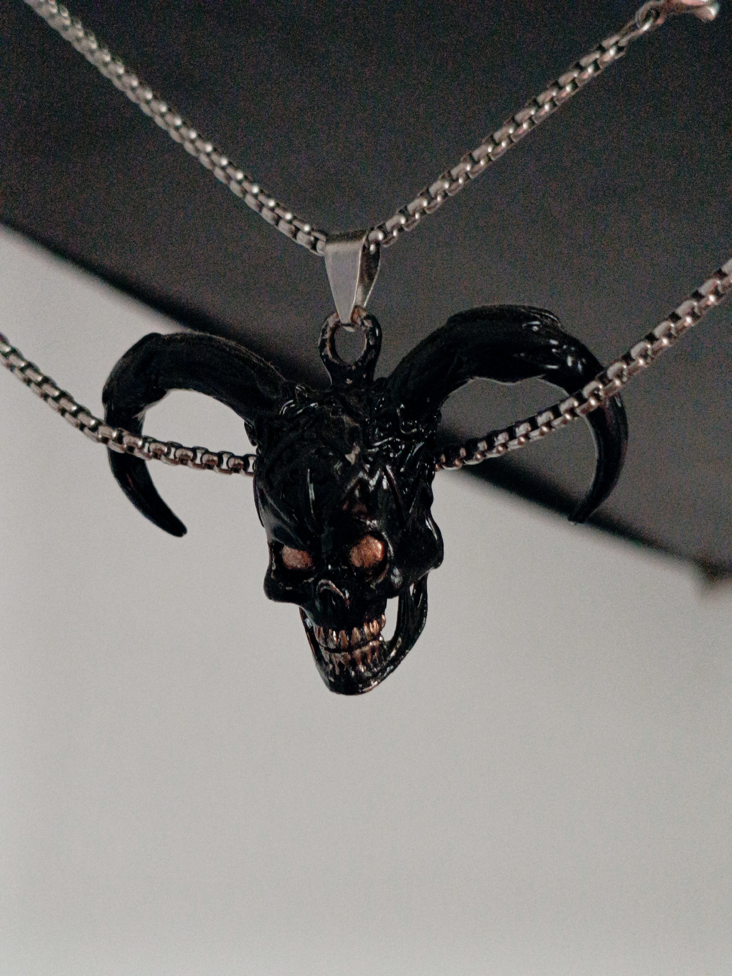 A close-up, slightly angled shot captures a detailed, black metal pendant shaped like a demonic skull with large, curved horns. The pendant is suspended from two separate silver-colored box chains, one draped above and the other below, meeting at a small silver clasp visible at the top center. The skull features hollow eye sockets with glowing orange or red irises, sharp teeth that are bared in a grin, and intricate detailing around the brow and cheekbones. The texture of the pendant appears slightly rough and metallic. The background is a gradient of dark gray at the top, transitioning to a lighter gray or white at the bottom. The image does not depict any specific urban environment or recognizable landmarks of Tash-Dobo, Kyrgyzstan.