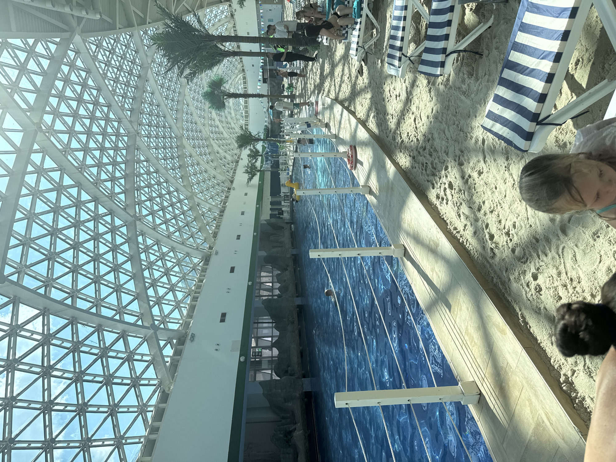 This image depicts the indoor wave pool at the Gazprom complex in Estosadok, Russia. The scene features a large, bright blue pool with rippling water, bordered by a sandy "beach" area. Several lounge chairs with blue and white striped cushions are scattered on the sand. Palm trees are visible throughout the space, adding to the tropical ambiance. The entire area is covered by a geodesic dome-like structure with a metal framework and translucent panels, allowing natural light to filter in. In the water, a few people are swimming. On the sandy area, a group of people are standing and sitting, some appearing to be staff or other patrons. A child with blonde hair is in the foreground, looking down. The overall impression is one of a recreational water park.
