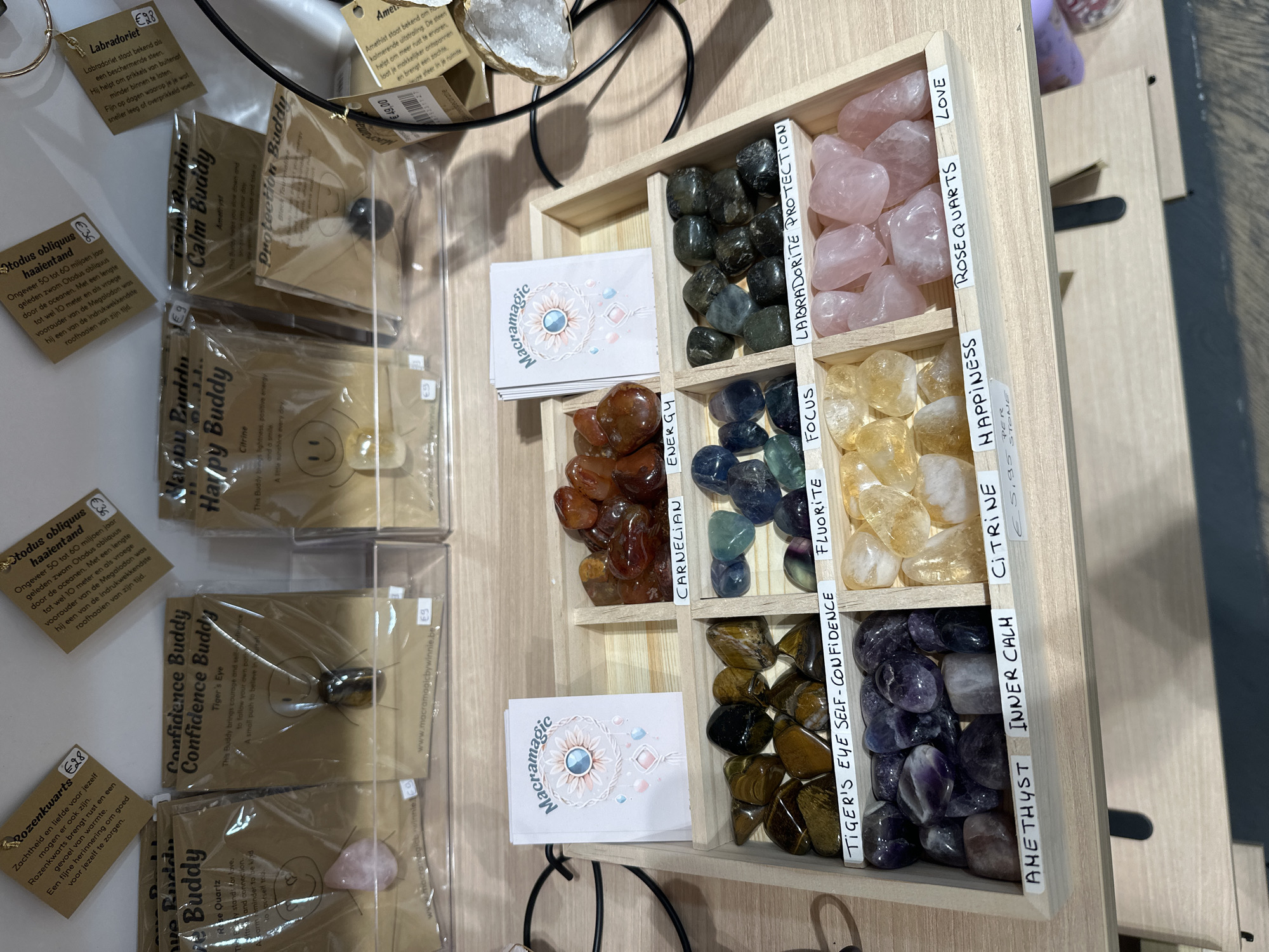 The image shows a retail display of polished stones and small charm pouches, likely in a shop in Bruges, Belgium. The stones are arranged in wooden trays and labeled with their names and supposed properties. For example, one tray contains Tiger's Eye stones labeled "Tiger's Eye Self-Confidence," another contains Amethyst labeled "Amethyst Inner Calm," and a third has Rose Quartz labeled "Rose Quartz Love." The charm pouches, also on display, appear to be small bags with stones or other decorative items inside, each with a card detailing its name and purpose, such as "Confidence Buddy" or "Calm Buddy." Some of these pouches have prices marked on them. In the background, there are more products, including what appears to be a large geode on a stand. The overall setting suggests a gift shop or a store specializing in crystals and wellness products.
