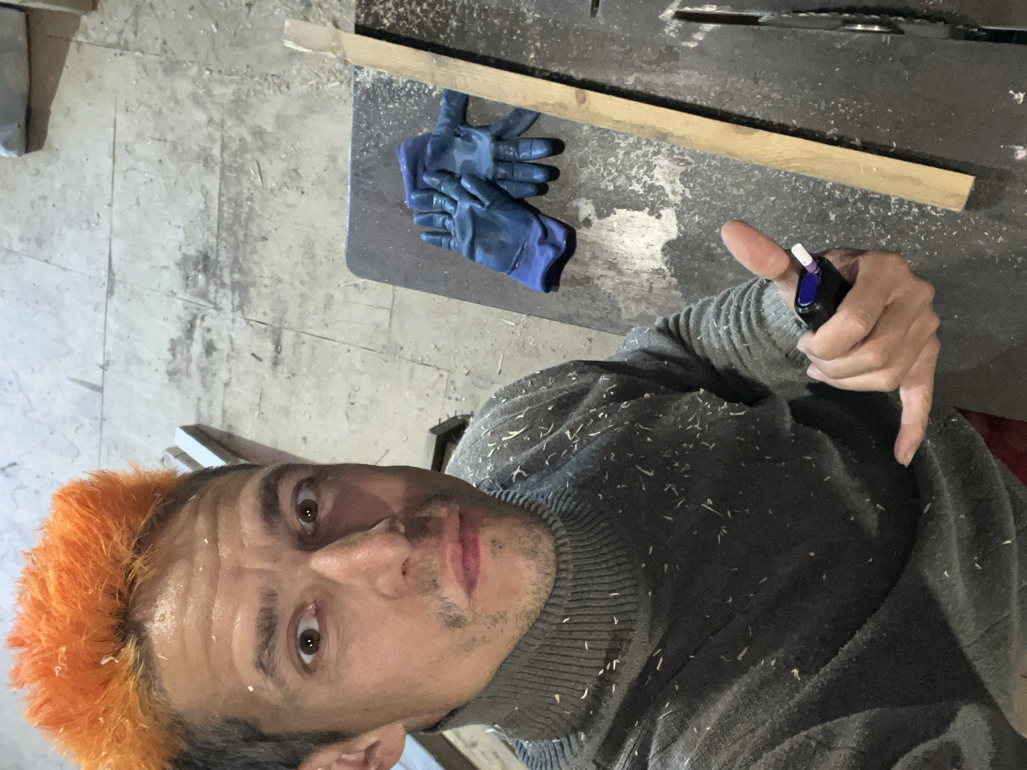 The image features a man in what appears to be a workshop or construction setting. He is looking directly at the camera, possibly taking a selfie. He has distinctive, bright orange-colored hair, mixed with light-colored wood shavings. His face shows some smudges of dirt or dust. He is wearing a gray ribbed turtleneck sweater, which is heavily covered in wood shavings.
The man's right hand holds a small black and purple tool, possibly a marker or stylus, with his thumb extended upwards in a casual gesture. Behind him, to his right, is a dark-colored workbench or saw table, also dusted with wood shavings. A light-colored wooden plank rests horizontally on this surface. A pair of blue work gloves are placed on the workbench to the left of the plank.
The background shows a concrete or tiled floor that is dirty and dusty, consistent with a work environment. The presence of wood shavings on the man's hair and sweater, and on the workbench, suggests recent woodworking activity. No text is visible in the image. The scene conveys a moment during or after manual labor in Chaikovsky, Russia.
