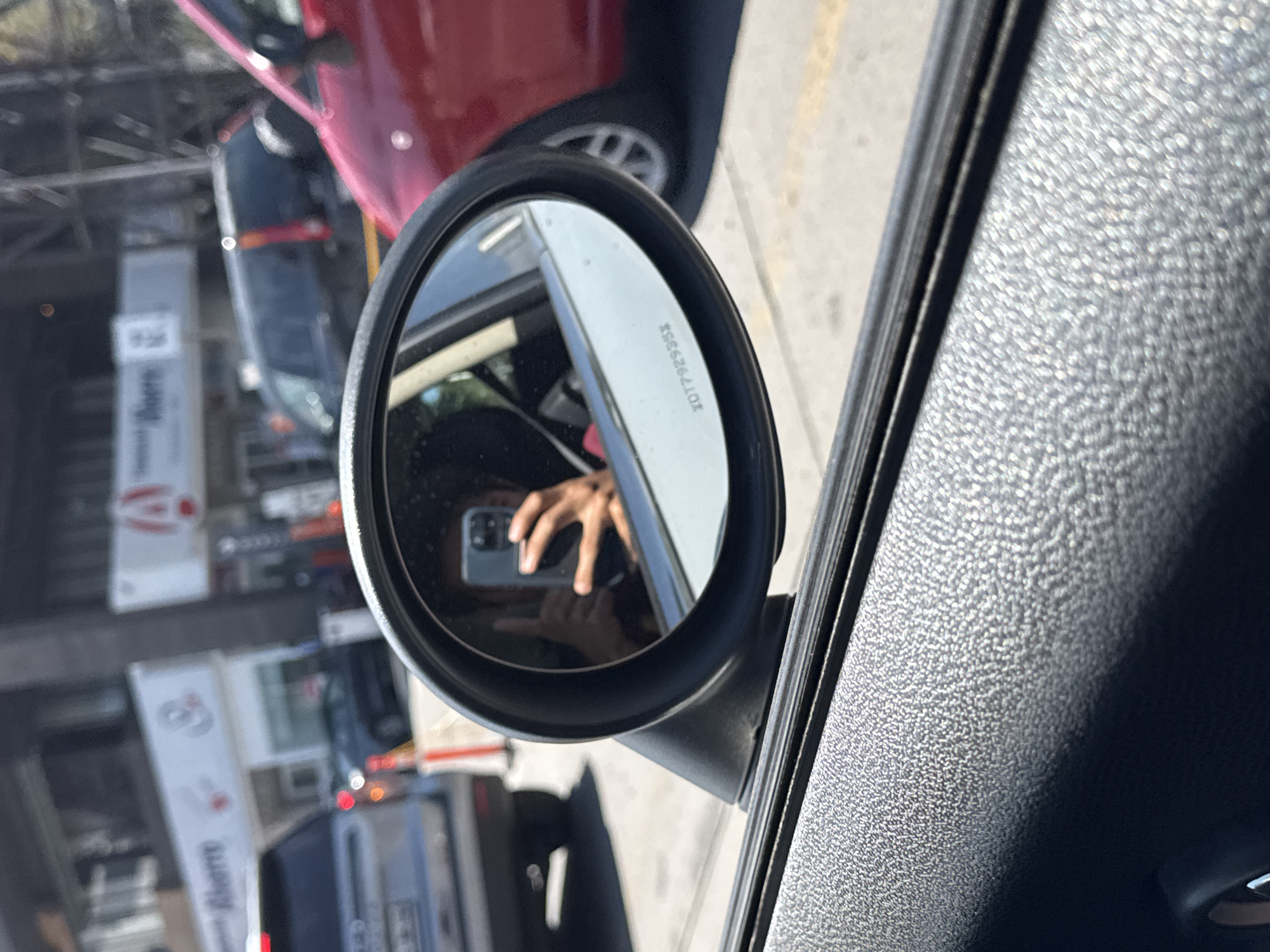 The image shows a driver's side mirror of a car, reflecting a person taking a photo of themselves with a mobile phone. The reflection also shows a street scene with a red car partially visible next to the mirror, and buildings with signage in the background. While the city is mentioned, there are no specific signs to confirm its location in San Bernardino Tlaxcalancingo, Mexico. The description is limited to what is directly visible in the image.
