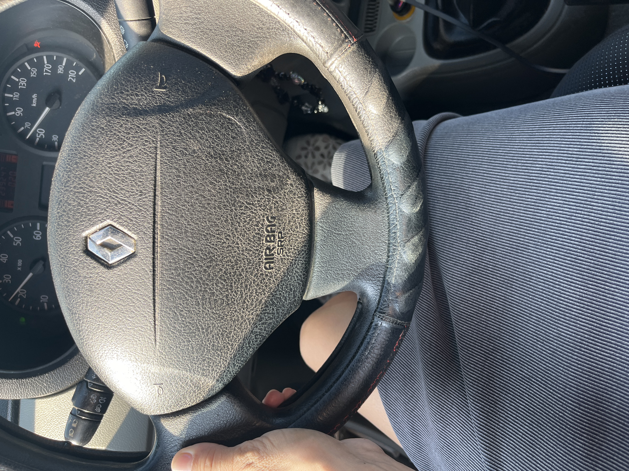 A person's hand is visible on the left side of the frame, grasping the steering wheel of a car. The steering wheel is black, textured, and features the Renault logo in the center. The dashboard includes a speedometer and other gauges. The person is wearing a gray, pinstriped skirt or shorts. The scene appears to be inside a car. The context of Mynai, Ukraine is not discernible from the image.