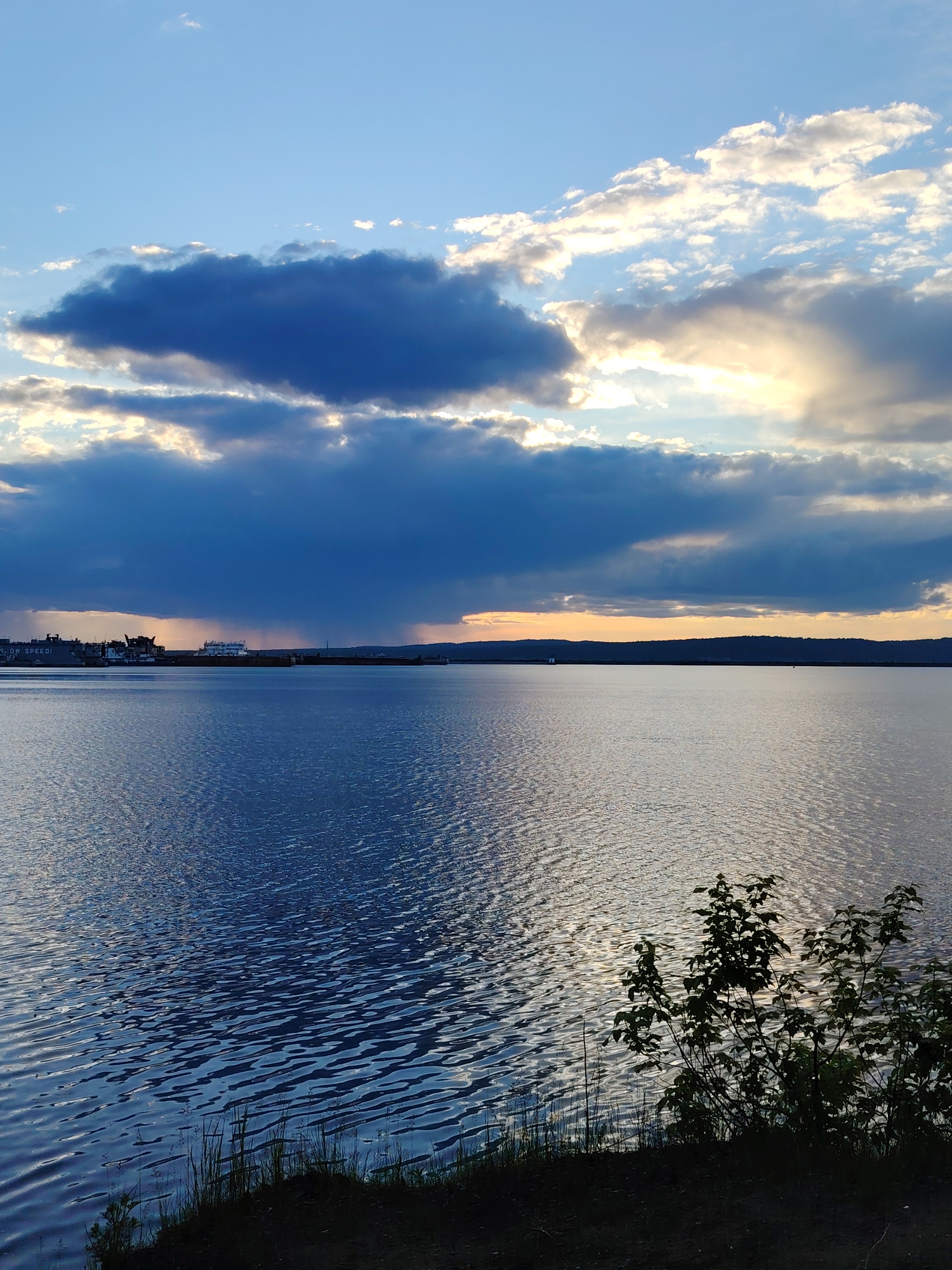 The image captures a body of water with ripples, likely a lake or river, under a sky with dramatic clouds. Some of the clouds are dark and heavy, potentially rain-bearing, while others allow sunlight to break through. In the distance, a town or industrial area with several ships is visible along the horizon. Closer to the viewer, vegetation is visible in the lower right corner of the frame. The location is Chaikovsky, Russia.