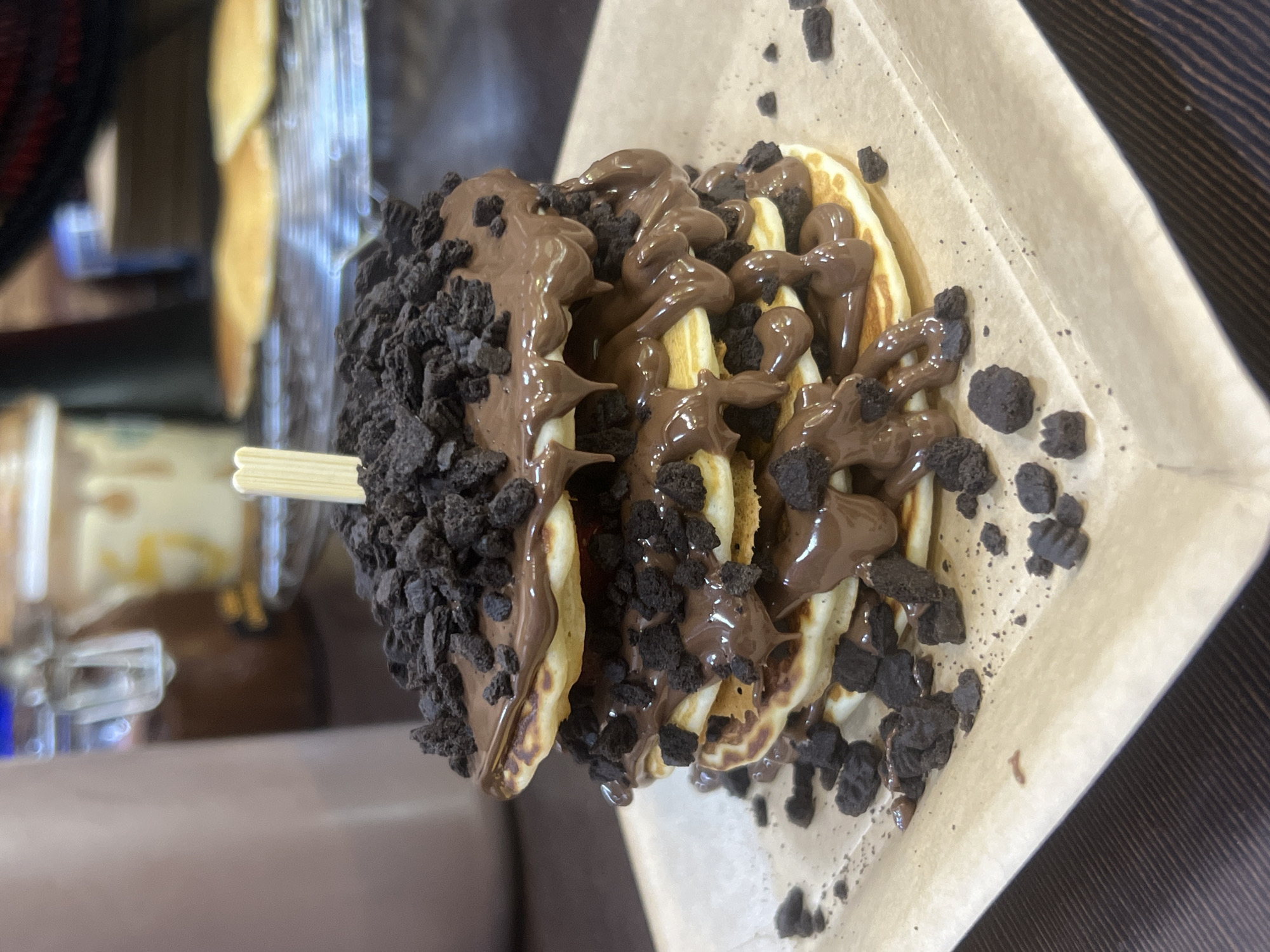 The image depicts a waffle on a stick, coated in chocolate sauce and crushed Oreo cookies, resting in a paper wrapper. It's likely a dessert item. The location, Plovdiv, Bulgaria, cannot be verified from the image content alone; therefore, I cannot determine the veracity of this claim. A blurred background shows hints of a cafe setting with other food items and equipment. The lighting is adequate, enabling clear visibility of the waffle and its toppings.