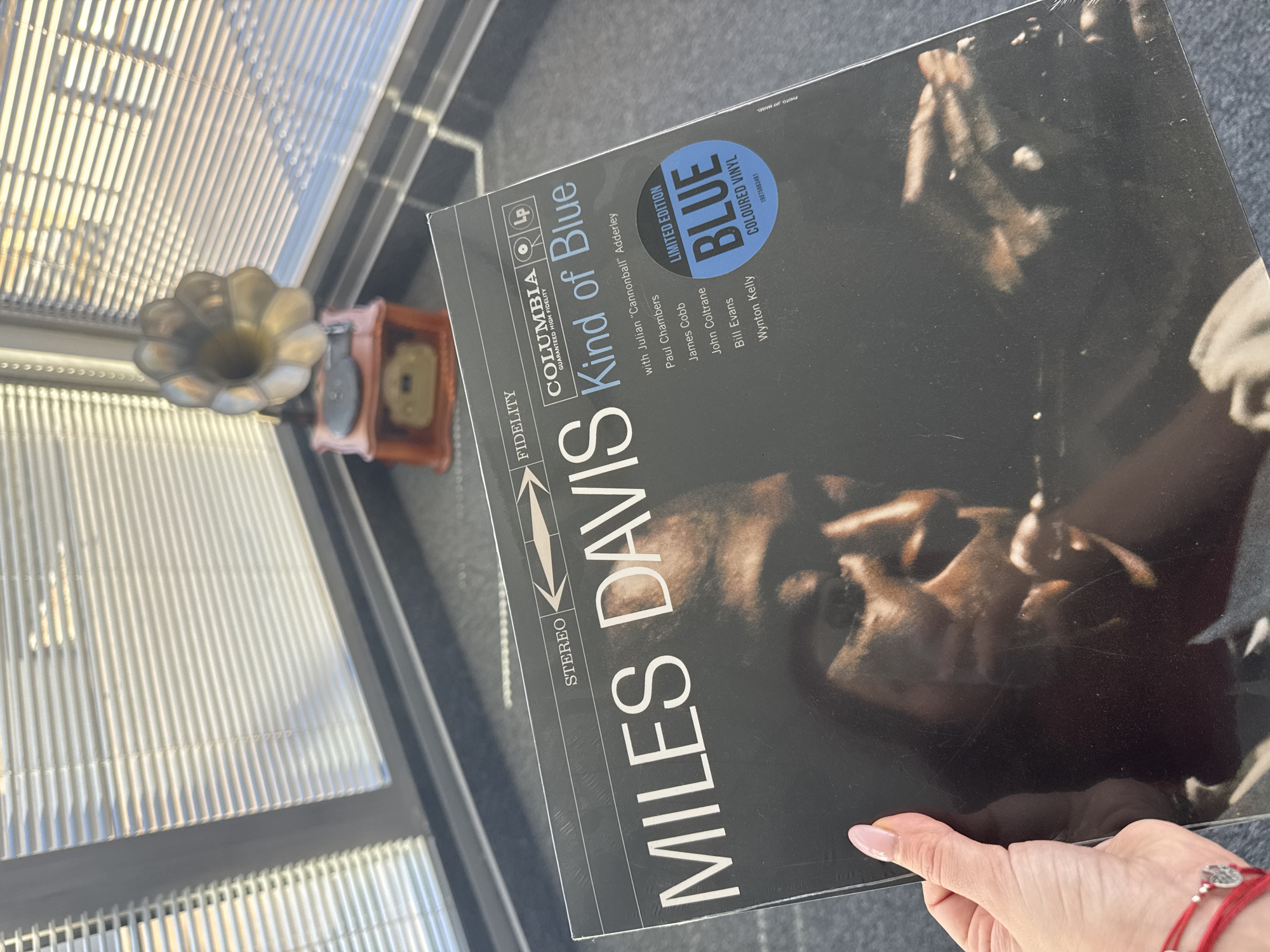 A hand holds a Miles Davis "Kind of Blue" vinyl record. The record jacket indicates a limited edition blue colored vinyl. A vintage gramophone sits atop what appears to be a window sill next to the record. Light is coming through the window, perhaps from outside. The location may be Sofia, Bulgaria, though there is no direct evidence in the picture confirming this.