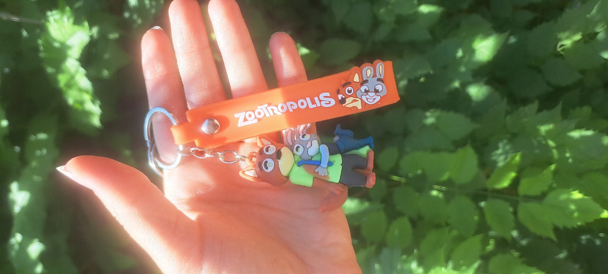 A close-up shot shows a hand holding a "Zootopia" themed keychain. The keychain features small figures of Nick Wilde and Judy Hopps, characters from the Disney movie, with Nick embracing Judy. The keychain also has an orange strap with "Zootopia" written on it in white lettering and additional character faces. The background is blurred green foliage, suggesting an outdoor setting in Bishkek City, Kyrgyzstan. The lighting indicates it is daytime, possibly sunny, with light casting on the hand and keychain.