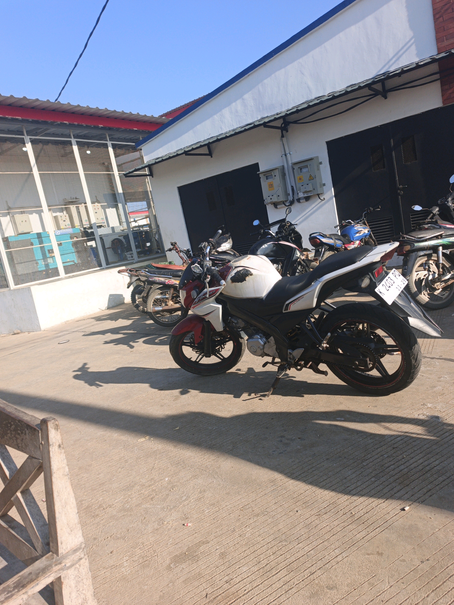 In the city of Unknown, Indonesia, on a sunny day, a row of motorcycles are parked in front of a building. The building has white walls and a red and blue roofline. There are two large black doors on the building, suggesting it may be a garage or workshop. Electrical boxes are mounted on the white wall. Several motorcycles are visible, including a prominent white and red one in the foreground with a license plate K 2433 PB. Other motorcycles, some with dark and blue color schemes, are parked behind it. The ground is a paved concrete surface with distinct lines, and shadows cast by the motorcycles and a wooden bench are visible. To the left, a building with large windows protected by mesh screens reveals equipment inside, possibly industrial machinery or electrical components. The overall scene depicts an outdoor parking area associated with a commercial or industrial establishment.