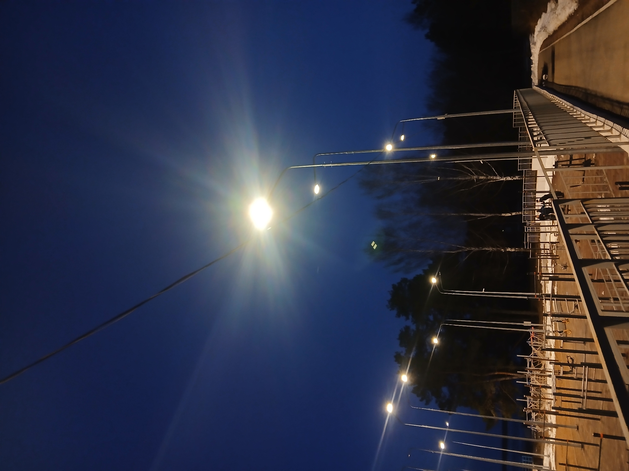 Here's a description of the image:
The image appears to be taken at night in what is possibly the city of Chaikovsky, Russia, based on visual cues. The perspective is angled upwards, capturing a footbridge or elevated walkway illuminated by a series of streetlights. The sky is a deep blue.
The bridge structure has metallic railings and a walking surface that appears to be concrete or similar material. The streetlights are tall and slender, with spherical luminaries that emit a bright glow, creating starburst effects in the image. Trees and other vegetation are visible in the background. There's evidence of snow along the side of the walking path.