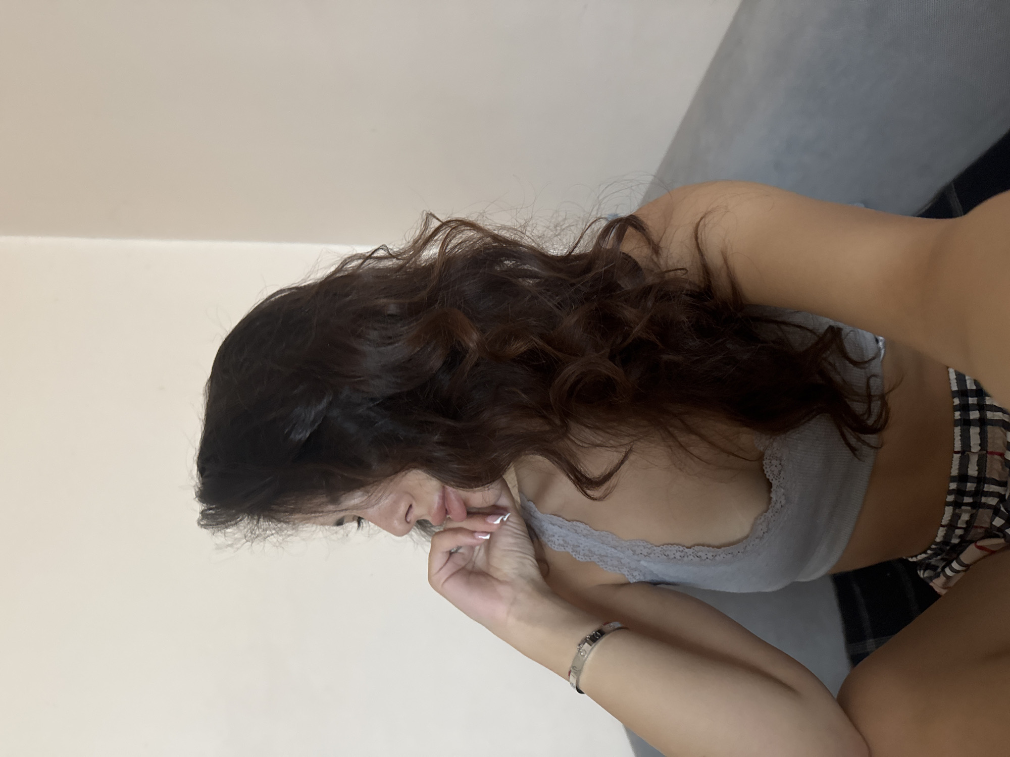The image shows a young woman with long, dark, wavy hair, wearing a light gray lace-trimmed bralette and plaid shorts. She is leaning forward, with her head bowed and her hand touching her mouth. Her fingernails are painted white. She is also wearing a silver bracelet on her left wrist. The background is a plain, light-colored wall and a gray sofa. The lighting appears to be artificial, suggesting an indoor setting, possibly in Plovdiv, Bulgaria. There are no specific time of day or weather cues visible. The woman's posture and hand gesture could suggest introspection or a moment of thought.
