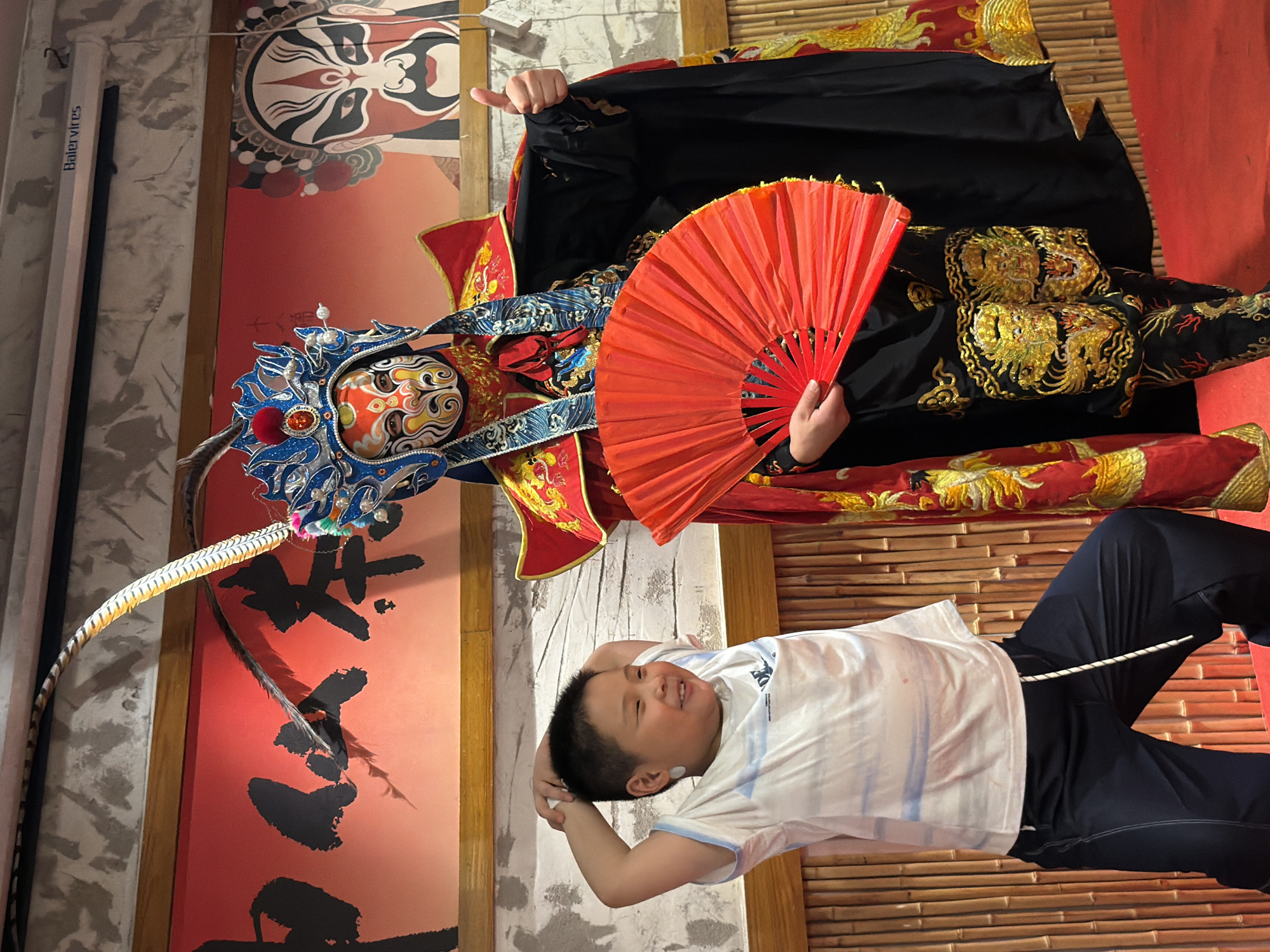 In Yuzhong District, China, a young boy with short black hair, wearing a white t-shirt with blue stripes and dark blue pants, poses with his hands behind his head, smiling. He is standing in front of a person in elaborate traditional Chinese opera attire, complete with a colorful mask, ornate headdress with feathers, a red and gold robe, and a black cape. This figure holds a vibrant red fan in one hand and gives a thumbs-up with the other. The background features a red banner with black Chinese calligraphy and a depiction of a traditional opera mask, with bamboo-like paneling to the left and a textured wall to the upper right.