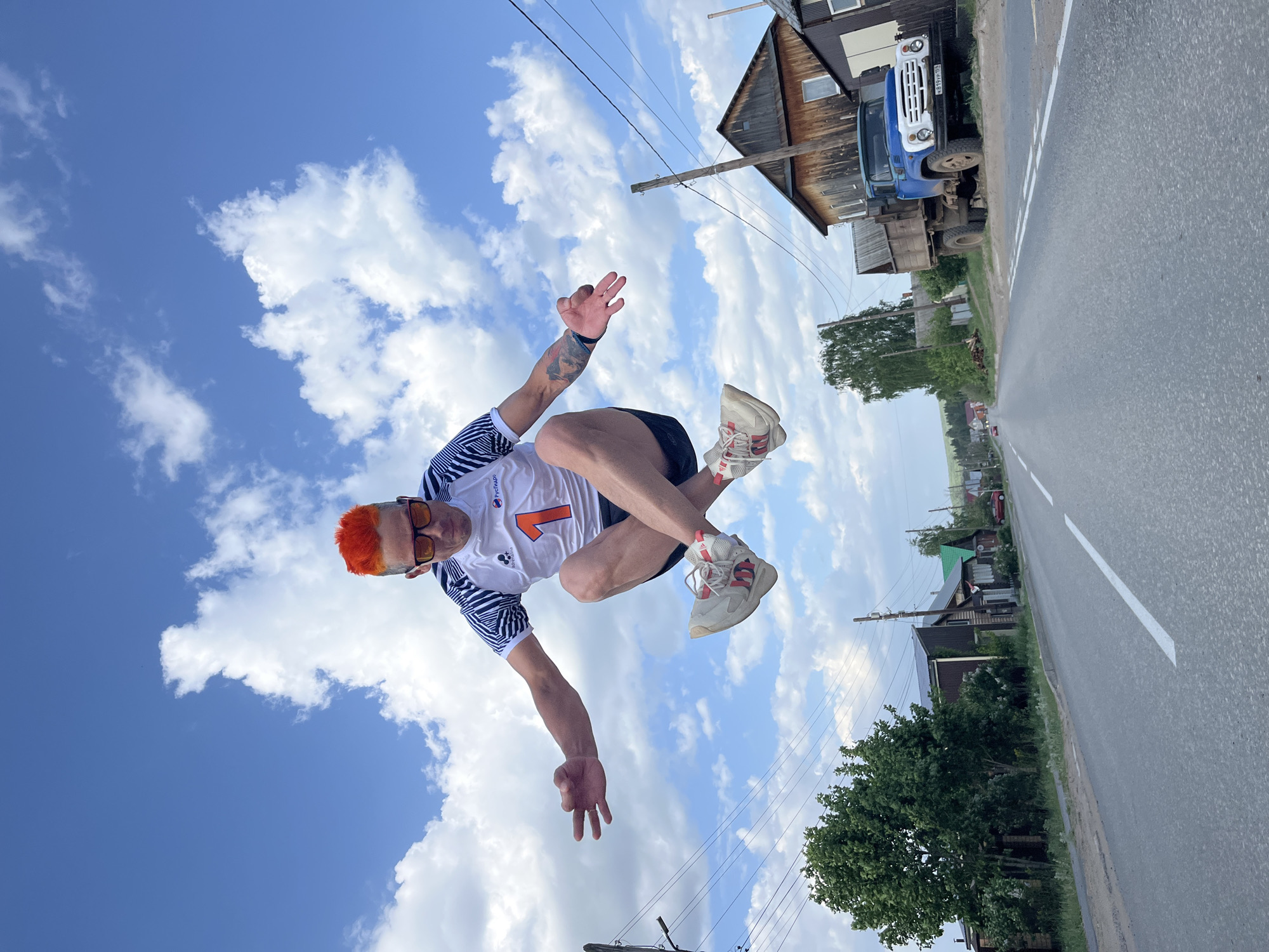 A man with orange hair and sunglasses is captured mid-jump on a road. He wears a white shirt with the number "1" and has tattoos on his left arm. A paved road runs horizontally across the frame, with a white dividing line. On the right side of the road are trees and residential houses. A blue truck is parked next to a wooden structure. Overhead, the sky is blue with scattered white clouds, and utility poles with wires stretch across the scene. The location appears to be a rural setting in the city of Chaikovsky, Russia.