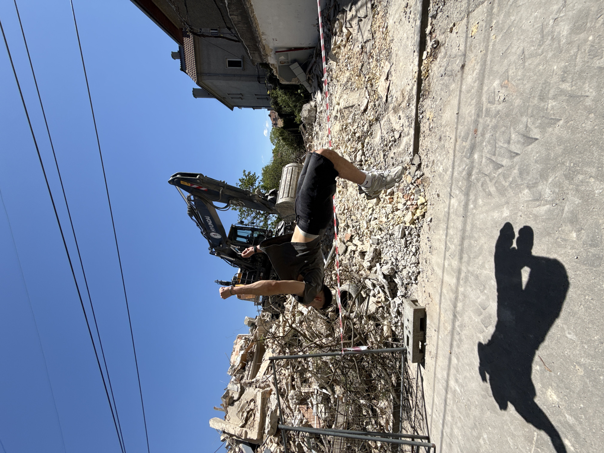 A man stands in front of the debris of a demolished building in Shumen, Bulgaria. He is wearing shorts, a t-shirt, and sneakers, and his arms are raised as if celebrating. Behind him is the demolished building, rubble scattered everywhere, a construction vehicle and a building can be seen in the background. Overhead power lines are visible against a clear blue sky. To the right, on a concrete surface, the shadows of two people, seemingly taking the photograph, are cast.