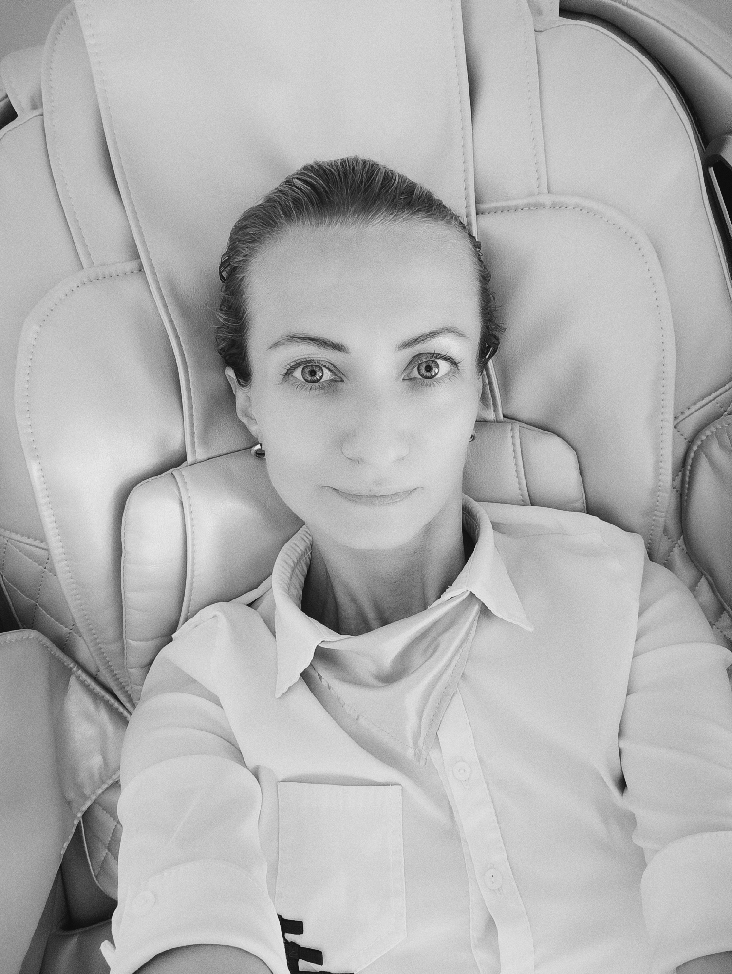 A black and white selfie shows a woman lying in a massage chair. She is wearing a white collared shirt and her hair is pulled back. The chair appears to be made of light-colored leather with a quilted pattern. The background consists of the textured upholstery of the chair. The image is taken from a high angle, looking down at the woman. The environment appears to be indoors. There is no visible indication of the location being Moscow, Russia.