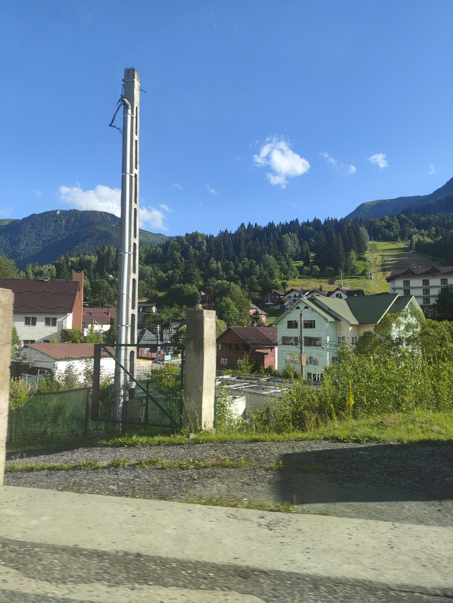 This image captures a view of the city of Borșa, Romania, on a clear day with a bright blue sky. In the foreground, a large concrete utility pole stands prominently, with some wires attached to it. To the left and right of the pole, partially obscured by greenery, are concrete pillars, suggesting a boundary or entrance. A dark metal fence is visible behind the utility pole and pillars.
The mid-ground of the image reveals a residential area with several houses. One house on the left has a reddish-brown roof and light-colored walls. Adjacent to it is another building with a similar roof and a prominent brick chimney. Further into the scene, there are more houses, some with green roofs, nestled amongst trees and vegetation. A ski lift is visible ascending a grassy slope in the background, indicating the area's connection to winter sports. The backdrop consists of lush green mountains covered in dense forest. The overall impression is of a semi-rural or suburban setting within a mountainous region.