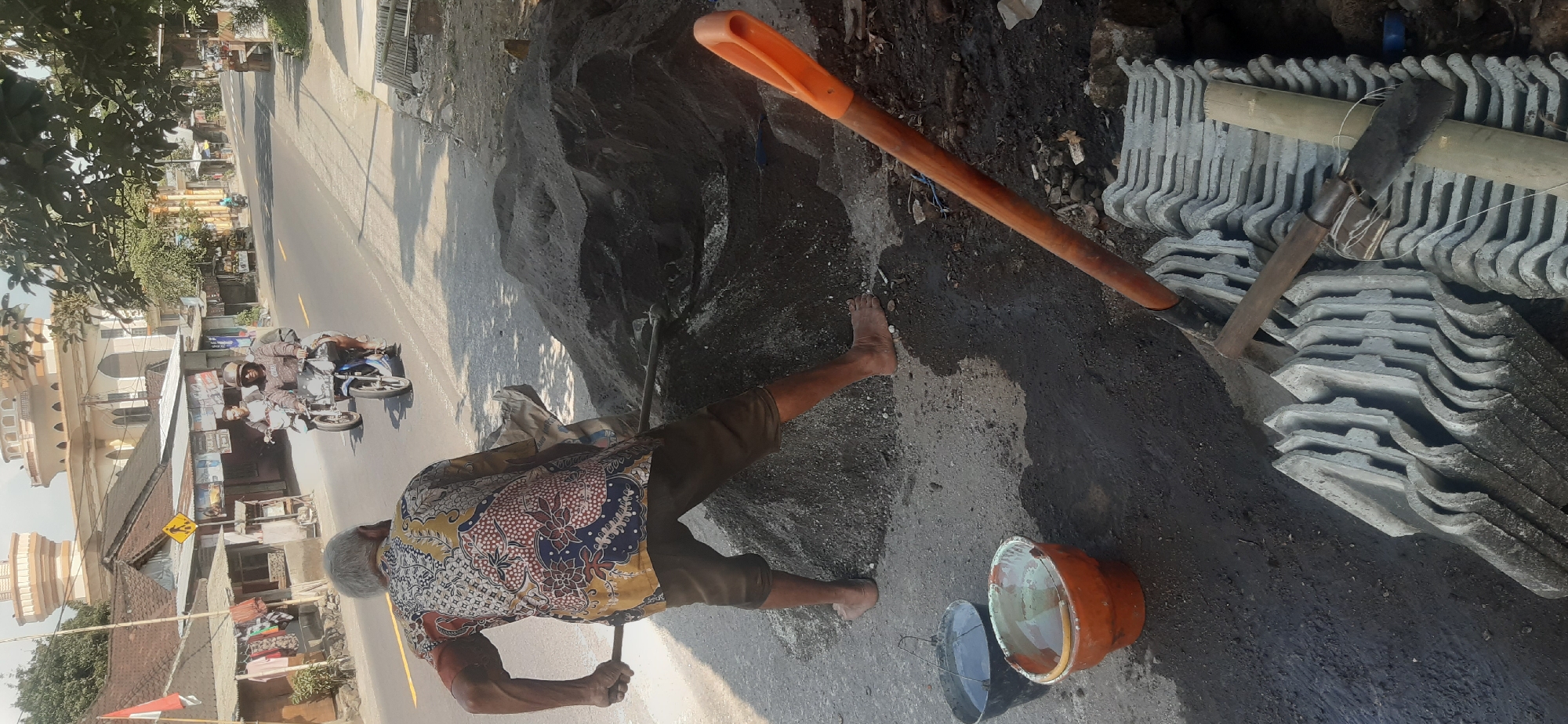 An elderly man with gray hair is in the foreground, wearing a colorful batik shirt and brown shorts, shoveling a pile of dark sand. He is barefoot. To his left, two buckets sit on the ground, one black and one orange, both containing water. A pile of gray roof tiles is stacked to the right of the sand pile, with two tools resting on them. In the middle ground, a road runs through what appears to be a village. Two men on a blue motorcycle are riding on the road. In the background, a white building with distinctive curved domes and arches, resembling a mosque, stands under a clear sky. The scene is outdoors with sunlight casting shadows. The overall impression is a work in progress in a semi-urban or village setting.