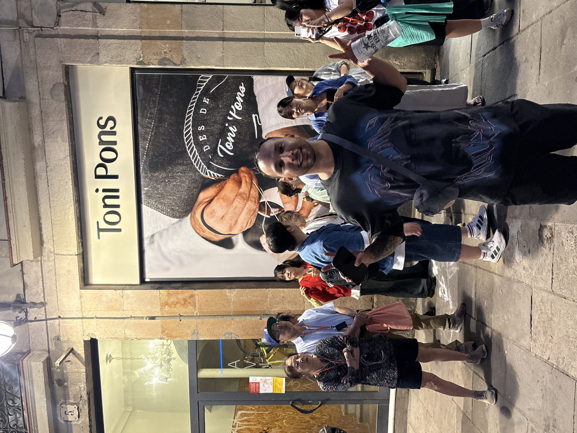 Here's a description of the image:
The photo shows a street scene in Barcelona, Spain, specifically in front of a Toni Pons store. A group of people stand on the sidewalk outside the store. The focus is on a man in the foreground, posing for the camera. The store's window display showcases an image related to the Toni Pons brand, featuring a person stitching espadrilles. Other details include the store's exterior lighting, a view of the interior with a chandelier, and a stone facade with the number 6 on a plaque. Several people in the background are tourists, either taking pictures or looking at the store.
