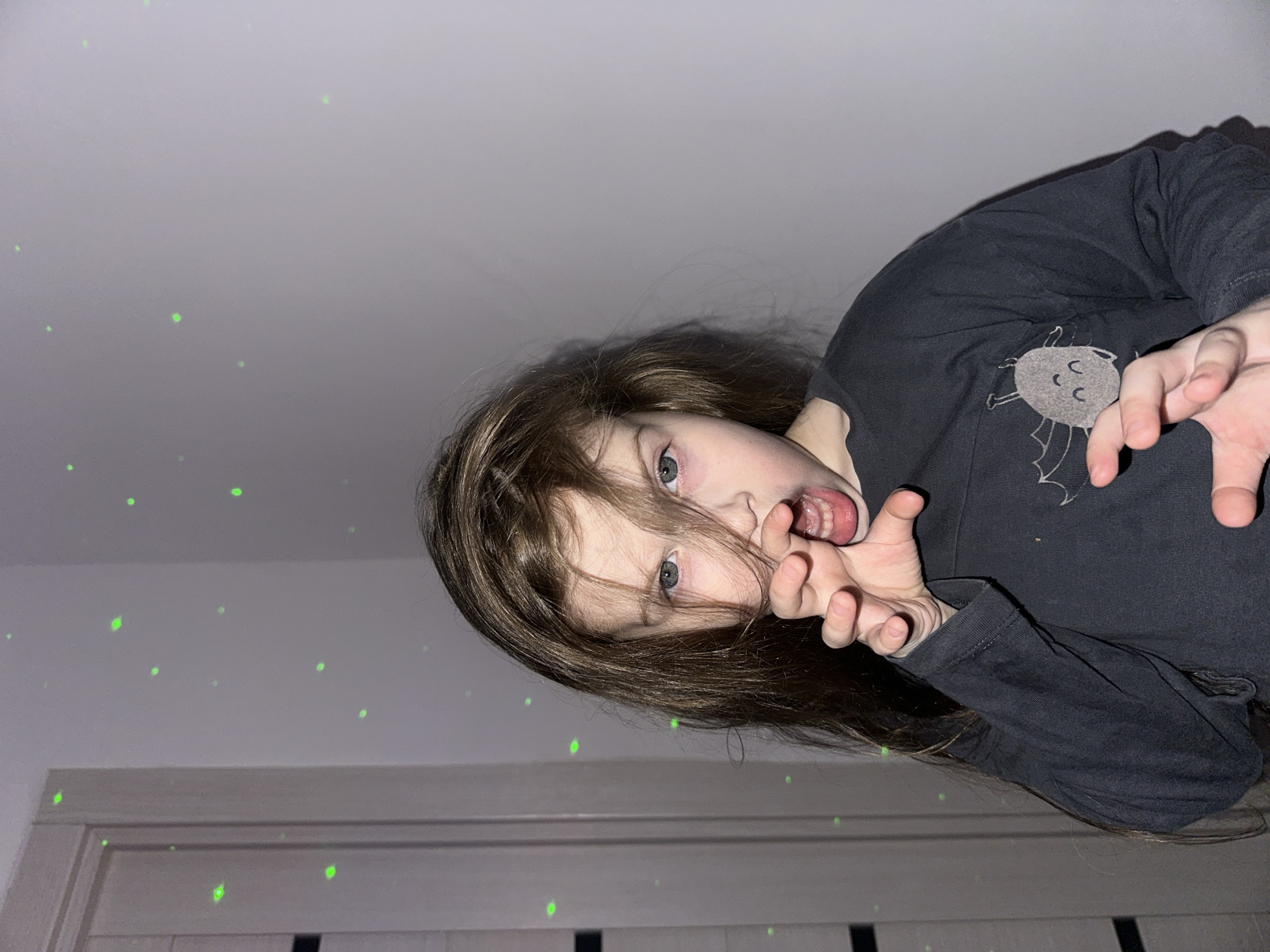 A child with long brown hair and light eyes wearing a dark long-sleeved shirt with a cartoon spider design is facing the camera. The child is making a playful, clawing gesture with their hands and has their mouth open in a pretended growl. The background consists of a white ceiling with scattered green laser dots and a white door frame. The location is Chaikovsky, Russia.