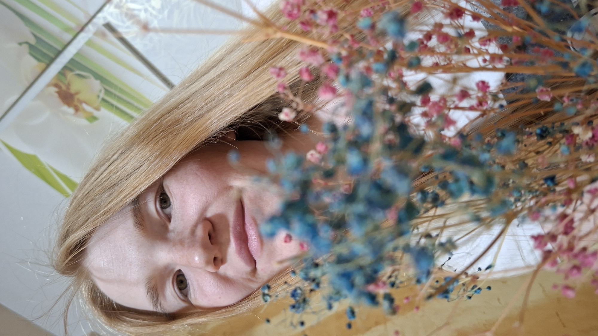 In an indoor setting, a young woman with blonde hair and light skin is captured in a close-up, angled shot. She is wearing a white garment. Her face is turned towards the camera, with her eyes looking directly at the viewer. Her lips are slightly parted, giving the impression of a soft smile or a sigh. She has a scattering of freckles across her nose and cheeks. The foreground is dominated by a cluster of dried flowers, a mix of vibrant blue and delicate pink, creating a soft-focus effect that blurs the background. The background appears to be a surface with a floral pattern, possibly wallpaper or a tablecloth. The overall lighting is soft and diffused, casting gentle shadows. The location is specified as Bolshoy Bukor, Russia, suggesting this scene is taking place within a residence in that area.