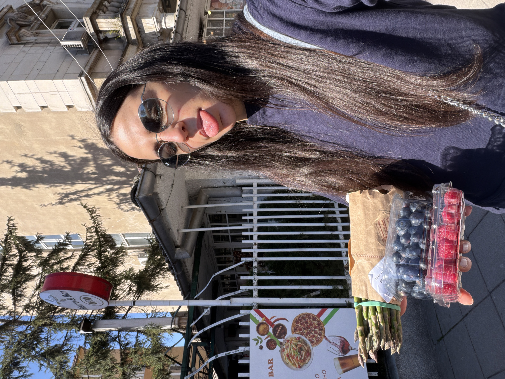 A young woman with long dark hair is seen outdoors, holding asparagus, blueberries, and raspberries. She is wearing sunglasses and sticking her tongue out. The background includes a building, a sign with a pizza and pasta advertisement, and another sign with "Bistro & Bar" written on it. The location appears to be in Sofia, Bulgaria, given the restaurant's advertisements are consistent with that location.