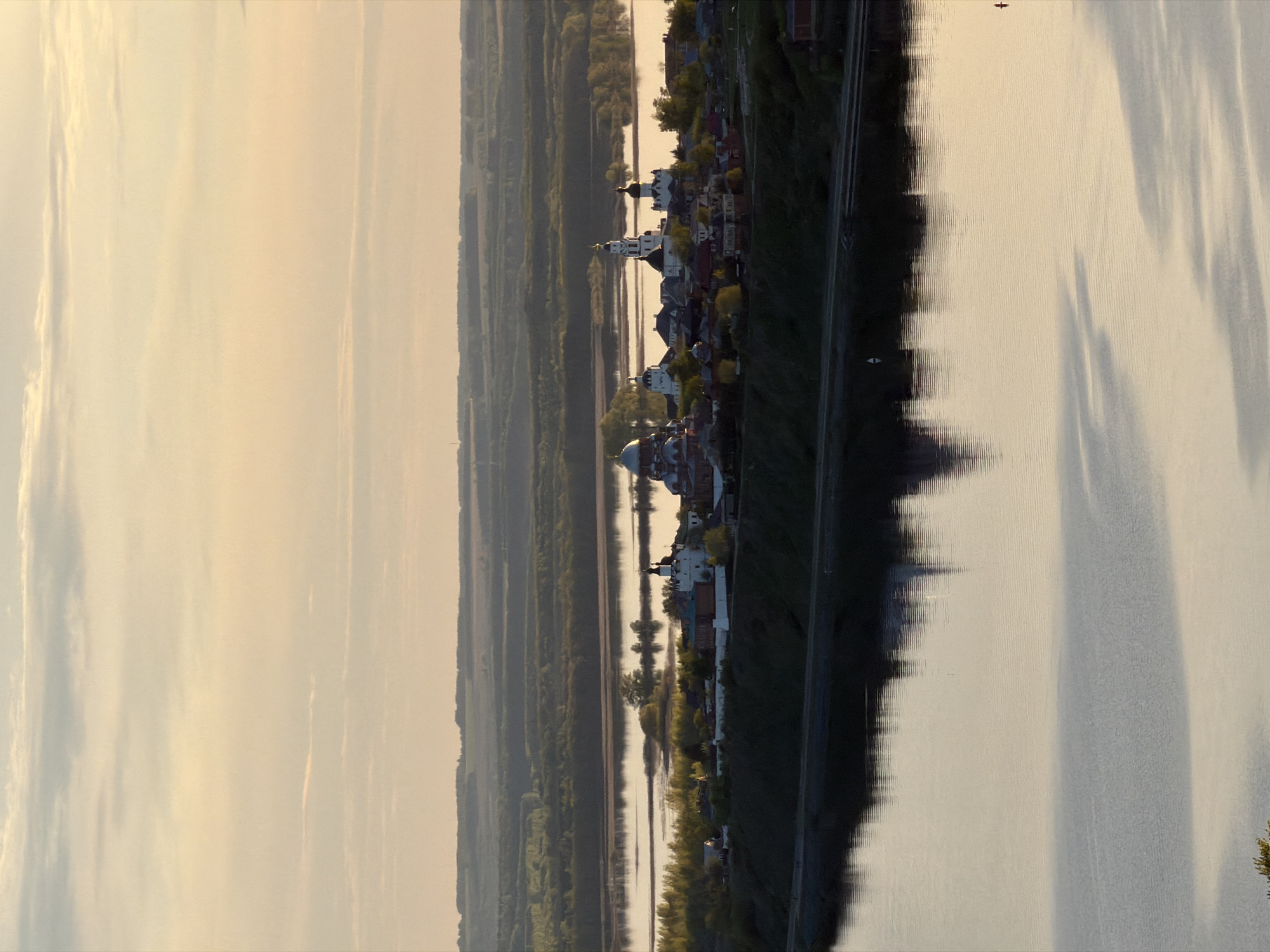 The image depicts a landscape view of Innopolis, Russia, likely taken from an elevated position. The scene showcases a river or a large body of water occupying a significant portion of the frame, with reflections of the sky and surrounding features visible on its surface. Along the riverbank, there is a row of buildings and structures, including what appears to be a cathedral with a prominent dome. The architecture suggests a historical or religious complex. Beyond the buildings, a dense forest stretches into the distance. The sky is filled with clouds, casting soft, diffused light on the landscape. The scene seems to be captured during the early morning or late afternoon, based on the warm, golden tones of the light.