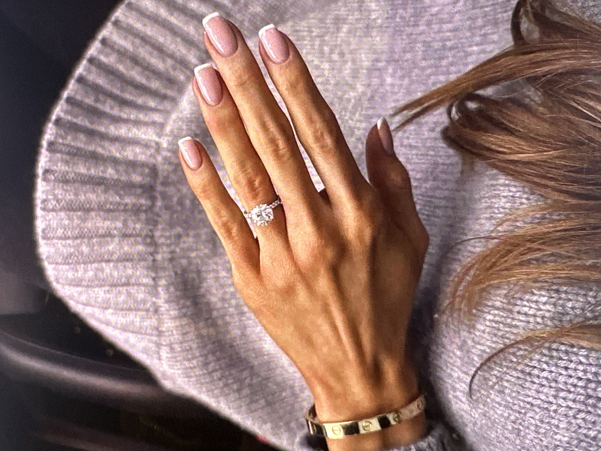 A woman's hand is prominently displayed, adorned with a large diamond ring on the ring finger and a gold Cartier Love bracelet on the wrist. The fingernails are manicured with a French tip design. The hand rests against a light gray, ribbed sweater. Brown hair is visible in the upper right of the frame. The image's location within Yerevan, Armenia, cannot be determined from the provided information.