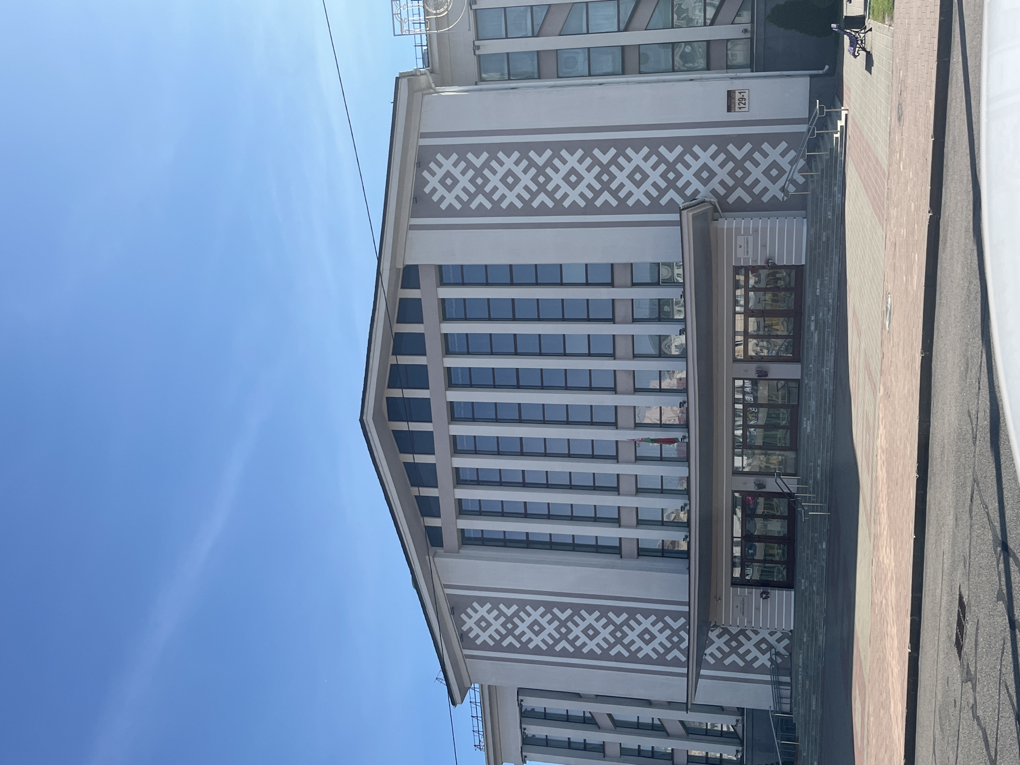 The image shows the exterior of a large, modern building with a light-colored facade, adorned with intricate, symmetrical geometric patterns in a darker shade on either side of the central section. The building has a prominent central entrance area with a wide staircase leading up to several glass doors. A flag, appearing to be the flag of Belarus, is visible flying near the entrance. The building's facade features a large number of tall, rectangular windows arranged in vertical sections. The sky above is clear and blue, indicating a sunny day. In the foreground, there is a paved area with brick-like patterns, and a street is visible in the lower part of the image. A small, dark-colored bench is situated on the right side of the paved area, and a trash receptacle is next to it. A sign with the number "129-1" is mounted on the building's facade to the right of the entrance. The building is likely a public or administrative facility in Minsk, Belarus.