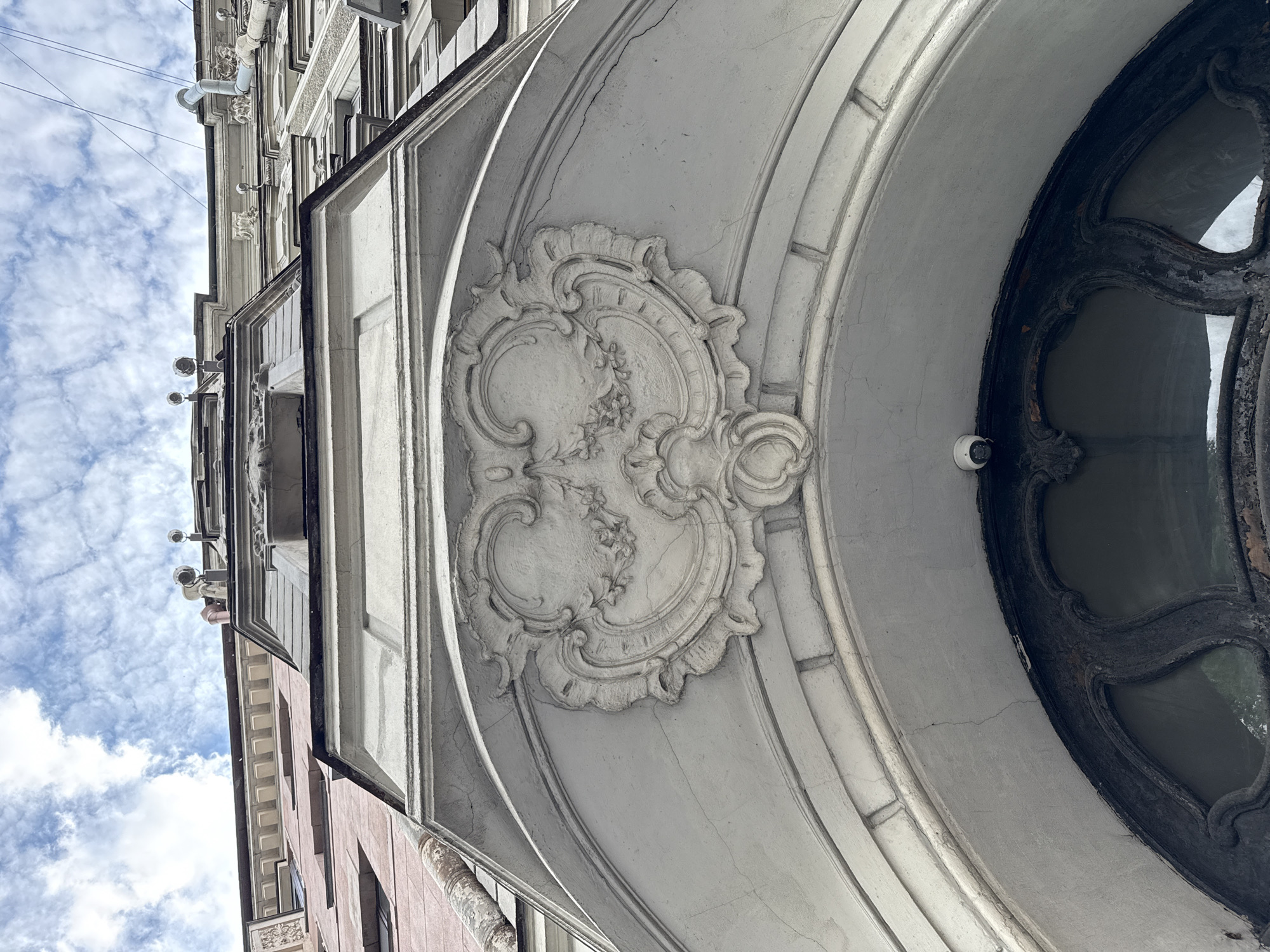 The image is a low-angle, close-up shot of the ornate facade of a building in Saint Petersburg, Russia. The focus is on an arched entryway featuring detailed stucco work. Above the arch, a decorative cartouche with swirling patterns and floral motifs is prominently displayed. The building's architecture appears to be in a classical or baroque style, with multiple levels visible receding upwards towards a cloudy sky. Various architectural elements, such as cornices, decorative moldings, and pipes, are present on the facade. A small security camera is mounted on the wall just above the arch. The overall impression is one of historical grandeur and architectural detail.