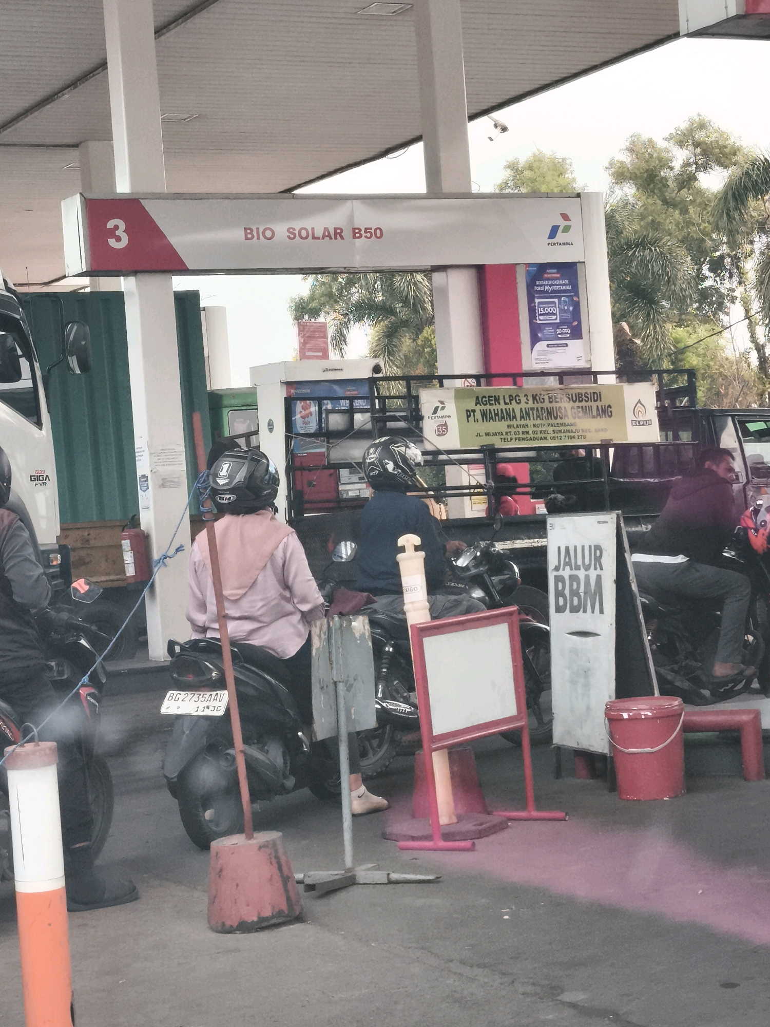 The image depicts a busy gas station in Palembang, Indonesia. At the forefront, two individuals are seated on motorcycles. One person, wearing a pink hijab and a light-colored top, is facing away from the camera, while the other, wearing a dark helmet and jacket, is partially visible from the side. Both are positioned at a gas pump.
In the background, a Pertamina branded gas pump displays "BIO SOLAR B50" and a number "3." Another pump area is visible with various containers and advertisements. A truck with a green metal side is parked to the left. A sign on a yellow banner advertises "AGEN LPG 3 KG BERSUBSIDI PT. WAHANA ANTARNUSA GEMILANG" with an address in Palembang and contact information.
Further to the right, a sign reads "JALUR BBM." A red bucket sits on the ground next to a red seating bench. The ground surface appears to be asphalt. Trees and foliage are visible in the background, indicating an outdoor setting. The overall scene suggests a typical day at a gas station with ongoing activity.