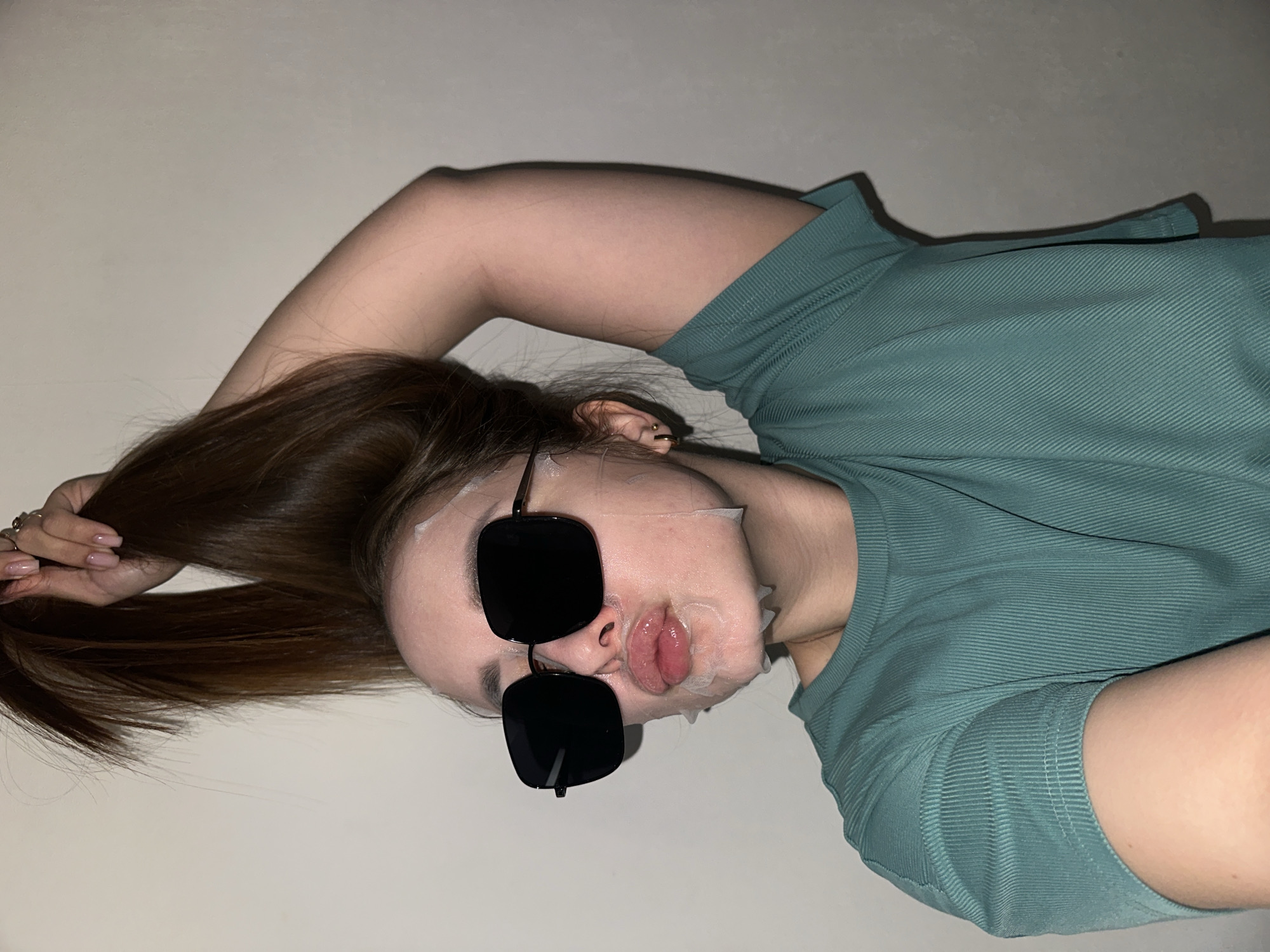 An informal, portrait-style photograph, rotated 90 degrees clockwise, captures a young woman from an overhead perspective. The primary subject is a woman with long, straight dark brown hair, which she holds up and out of the frame with her left hand. Her nails are manicured with a light pink or nude polish, and she wears a ring on one finger.
She is depicted wearing a light teal or mint green ribbed t-shirt or top. Her face is covered with a white sheet face mask, and she sports large, dark, square-framed sunglasses, covering her eyes. Her lips are puckered into a 'fish face' or kissy pose through the mask's mouth opening. A small ear piercing is visible on her left ear.
The background is a plain, light-colored wall, likely white or off-white, suggesting an indoor setting. The lighting appears soft and artificial, hinting at an evening or nighttime activity. The overall mood conveyed is relaxed, playful, and humorous, capturing a personal moment of self-care. There is no visible text in the image. This scene takes place in Chaikovsky, Russia.