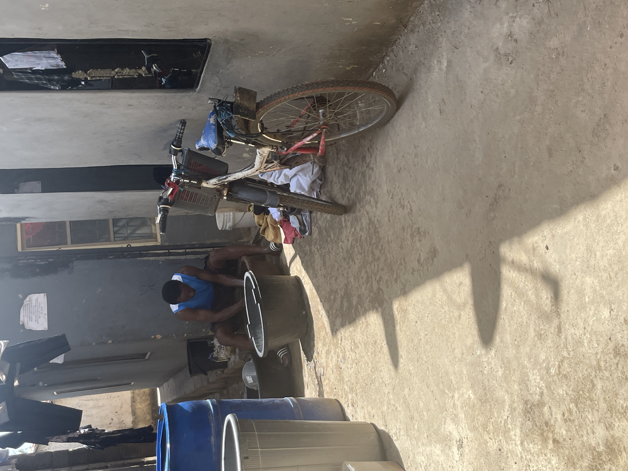 The image depicts a man sitting in front of a large, dark-colored cylindrical container, seemingly engaged in washing or some task related to the container. He is wearing a blue tank top with white trim and dark shorts. Beside him, also in front of the container, is a large, rusty-red bicycle, leaning against a wall. Clothes are draped over the bicycle and in the background. The scene is outdoors, with a concrete or dusty ground surface. In the background, there are buildings with windows and what appear to be other large plastic containers, some blue, lined up. The sun is casting strong shadows, suggesting it is likely daytime, possibly mid-morning or afternoon. The overall impression is of a simple, possibly utilitarian, outdoor space. There is no discernible text visible in the image.