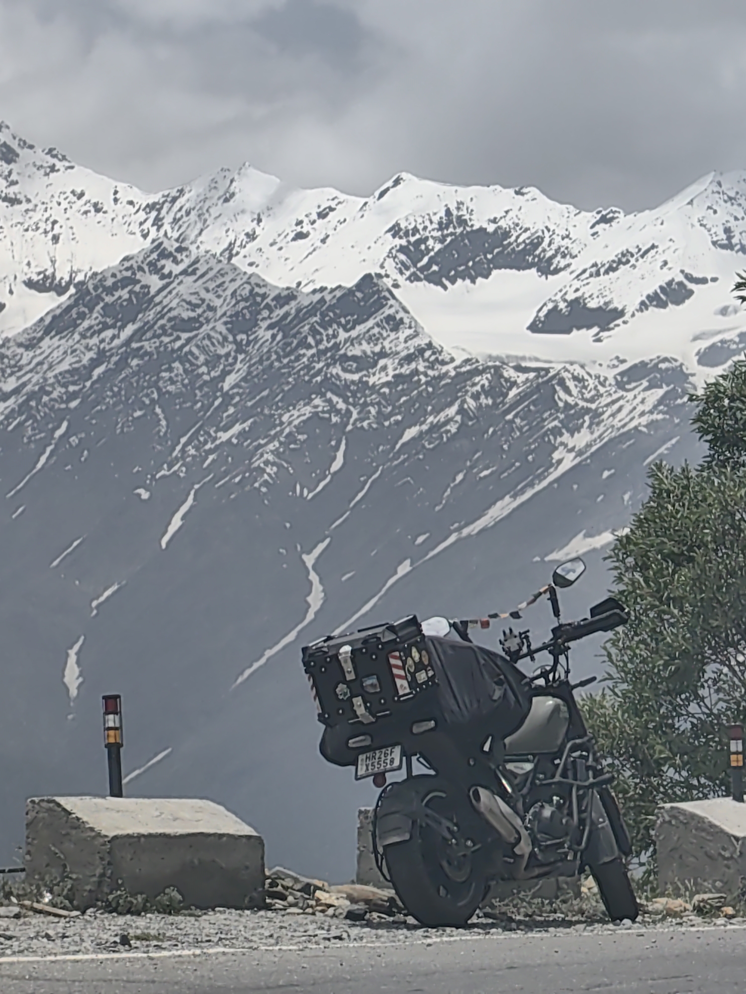 A motorcycle is parked on the side of a road with a backdrop of snow-capped mountains and a cloudy sky. The motorcycle has a large black travel case on the back, adorned with several stickers, and a smaller case is visible on the side. A license plate with the inscription "HR26F X5558" is attached to the rear fender. Prayer flags are strung across the handlebars of the motorcycle. In the foreground, to the left, a concrete block with a traffic marker featuring red, white, and yellow bands is situated. To the right, a lush green bush grows beside another concrete barrier. The ground is covered with gravel and debris. The mountainous terrain appears to be a natural landscape, possibly a scenic route or viewpoint in the region of Kinnaur, Himachal Pradesh, India, which encompasses areas like Yamling.