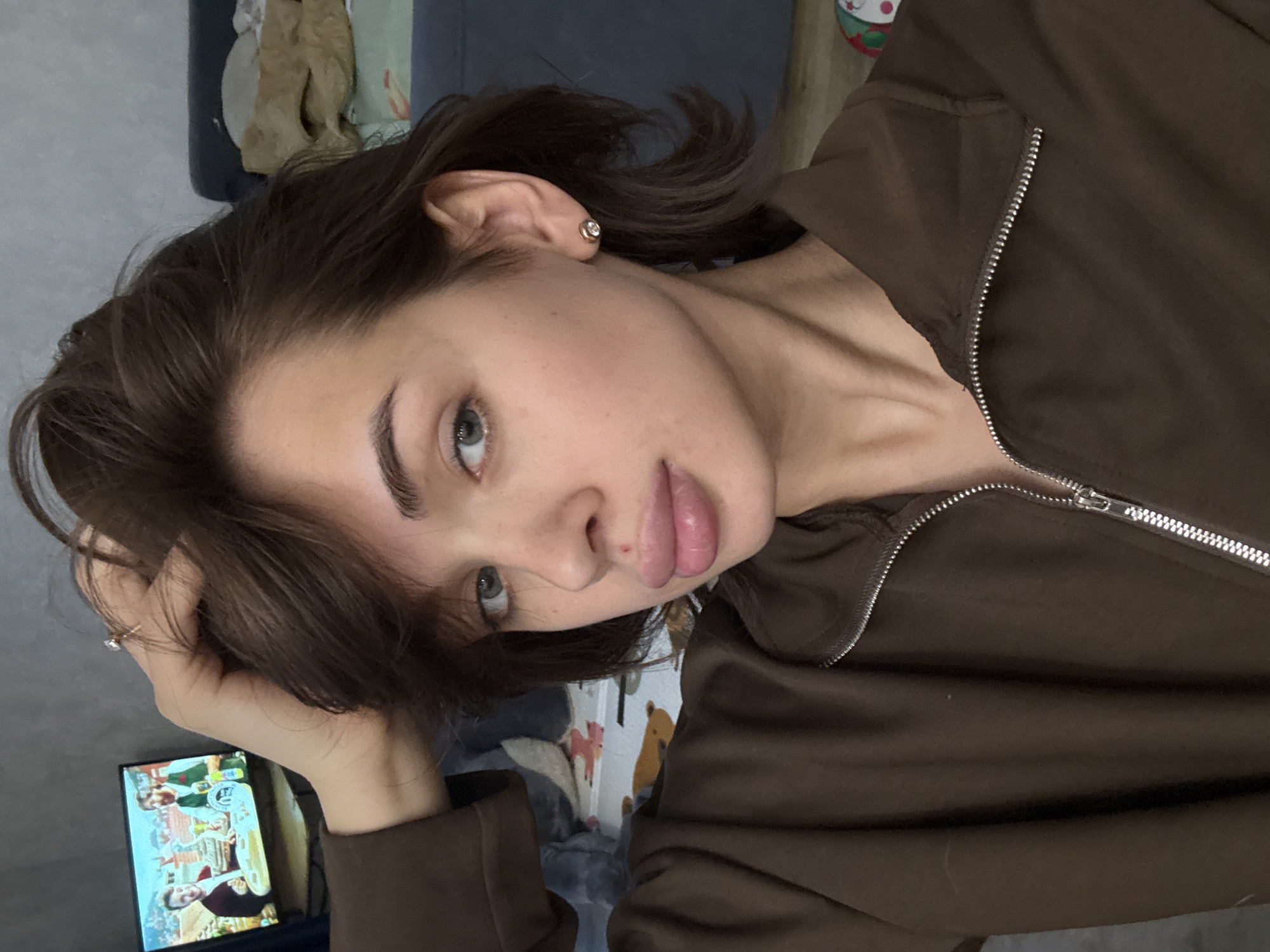 A close-up, angled selfie of a young woman. She has brown hair styled with some strands falling across her face, light-colored eyes, and full lips. She is wearing a brown zip-up jacket. She has an earring on her left ear. The background is blurry, but there is a TV screen showing people, a bed with various covers, and what appears to be a wall with cartoon-like illustrations. There is no indication of the location being Лобаново, Russia.