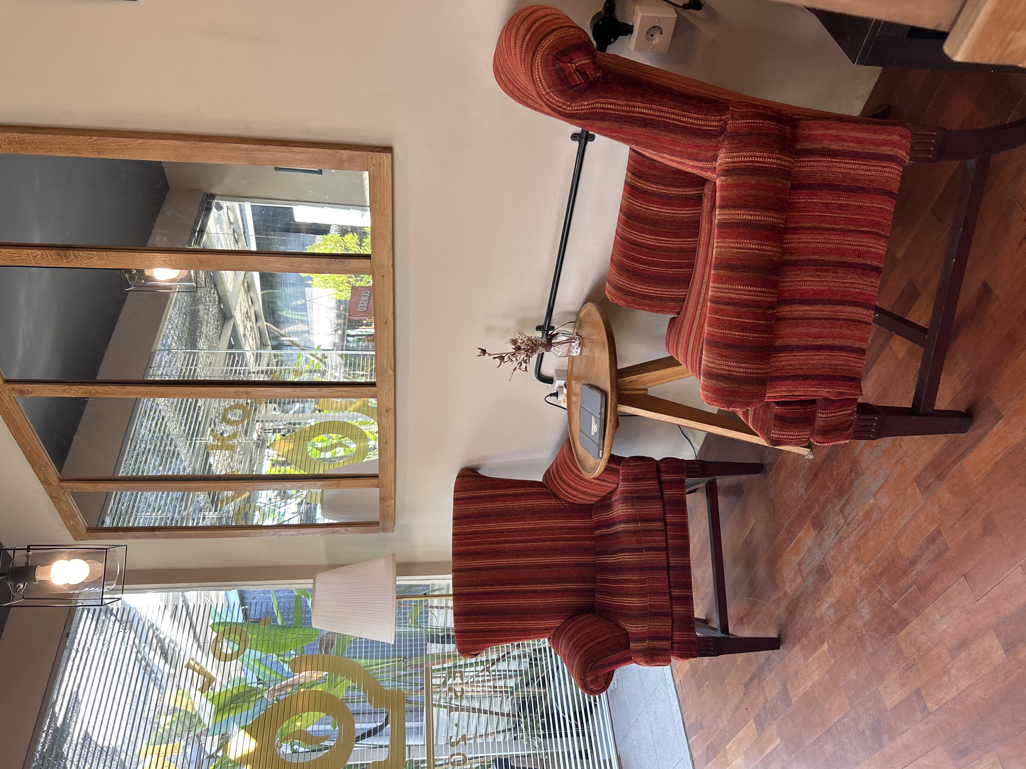 In a room with light brown walls and a wooden floor, two red striped armchairs are positioned. The armchair on the left has a white lamp with a pleated shade next to it. Between the armchairs is a small round wooden table with a tablet and a small vase containing dried flowers. A horizontal black bar is mounted on the wall behind the table, and an electrical outlet is visible to the right of the right armchair. A large wooden-framed mirror with multiple panels hangs on the wall above the armchairs, reflecting the interior. Through the blinds and window to the left, greenery is visible, along with a logo on the window. The scene suggests a comfortable seating area within a building, possibly a cafe or lounge, located in North Kuta, Indonesia, judging by the context of the request.