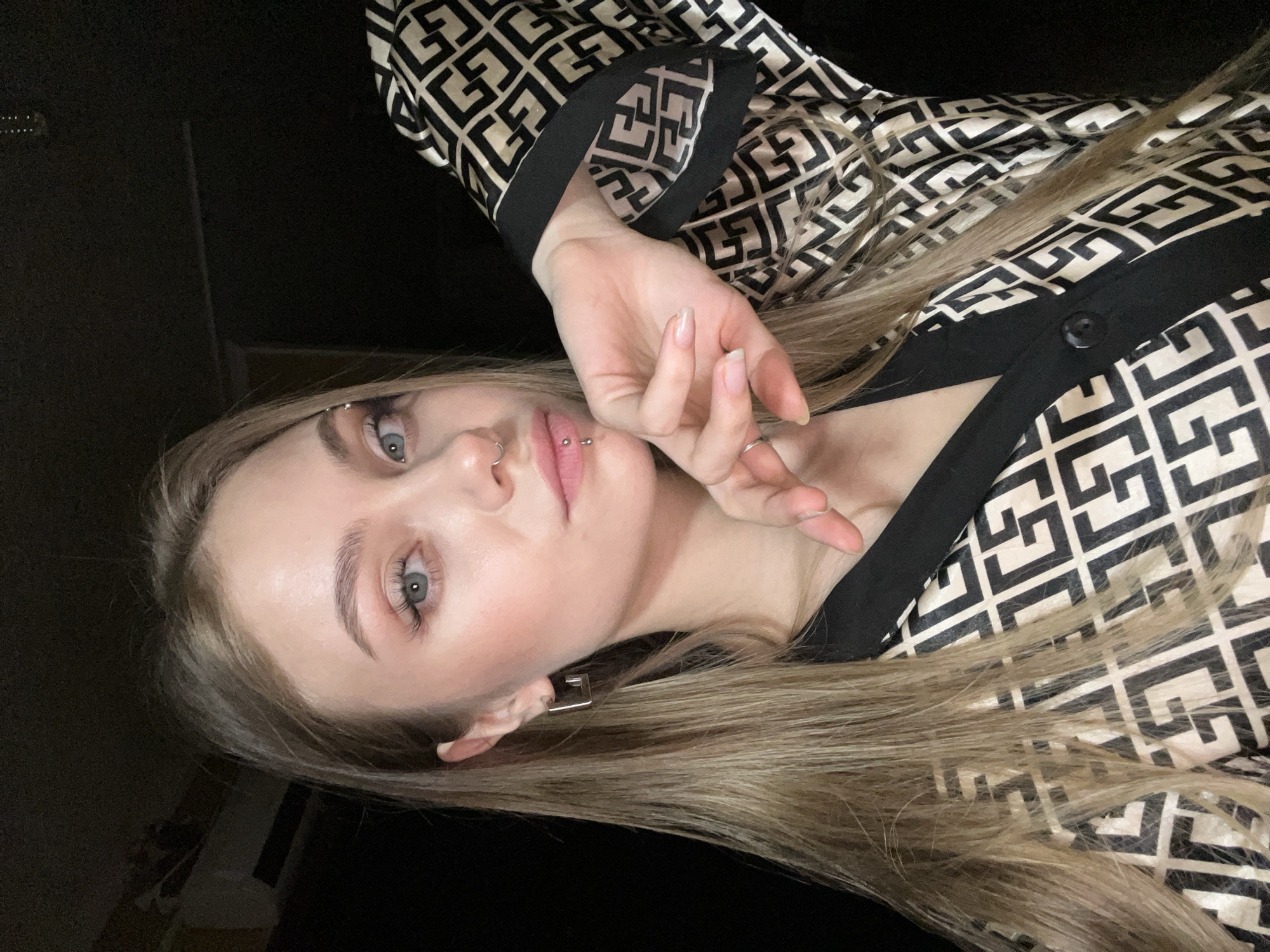 A young woman with blonde hair and fair skin is posing indoors, oriented at an angle relative to the camera. She has light blue eyes, pierced eyebrows, and lip piercings. Her right hand is raised near her face. She wears a black and beige patterned top with a black collar and a single button visible. The background is mostly dark and indistinct. The location, Фоки, Russia, is not discernable from the visual information.