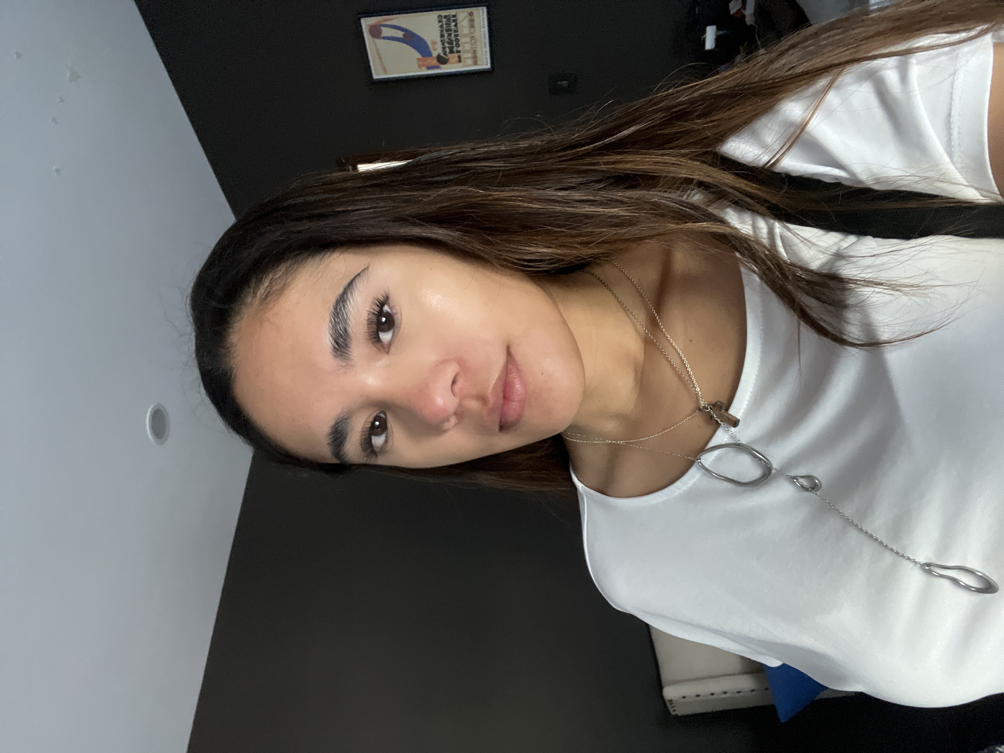 A woman with brown hair is facing the camera. She is wearing a white shirt and a silver necklace. The background is a dark gray wall, and a framed picture hangs on the wall. The location is unknown and cannot be determined based on the image.