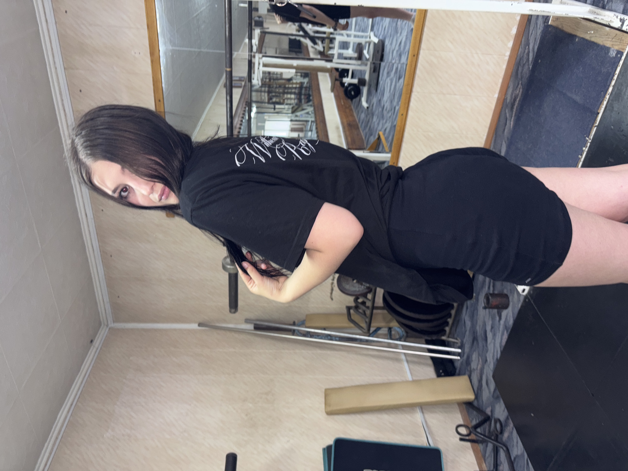 A young woman with long dark hair is posing in a gym. She is wearing a black t-shirt and black shorts. She is standing sideways to the camera, looking over her shoulder. Her left hand is holding her hair. The gym appears to be a home gym or a small, possibly older, commercial gym, with weights, weight racks, and a mirror. The walls are paneled and the floor is carpeted. There are no explicit indicators of the specific location within the city of Chaikovsky, Russia, but it is a gym setting.