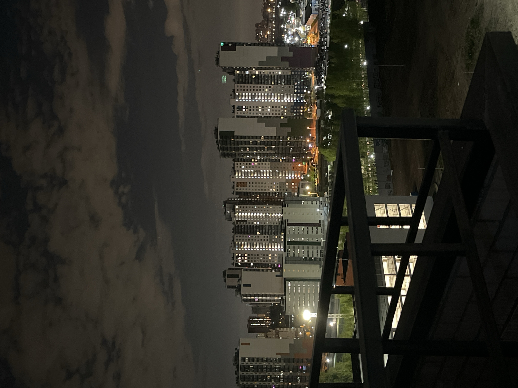 Captured from an elevated vantage point, the image presents a cityscape at night. The focus is on a series of high-rise buildings with numerous illuminated windows, suggesting residential or commercial occupancy. A dark, potentially cloudy sky forms the backdrop to the left of the frame. To the right, a dark, partially obscured structure, possibly a balcony railing, frames the view. Further in the distance, a strip of greenery and streetlights line the edge of the buildings. The specific city and location are not discernable from the visual information.