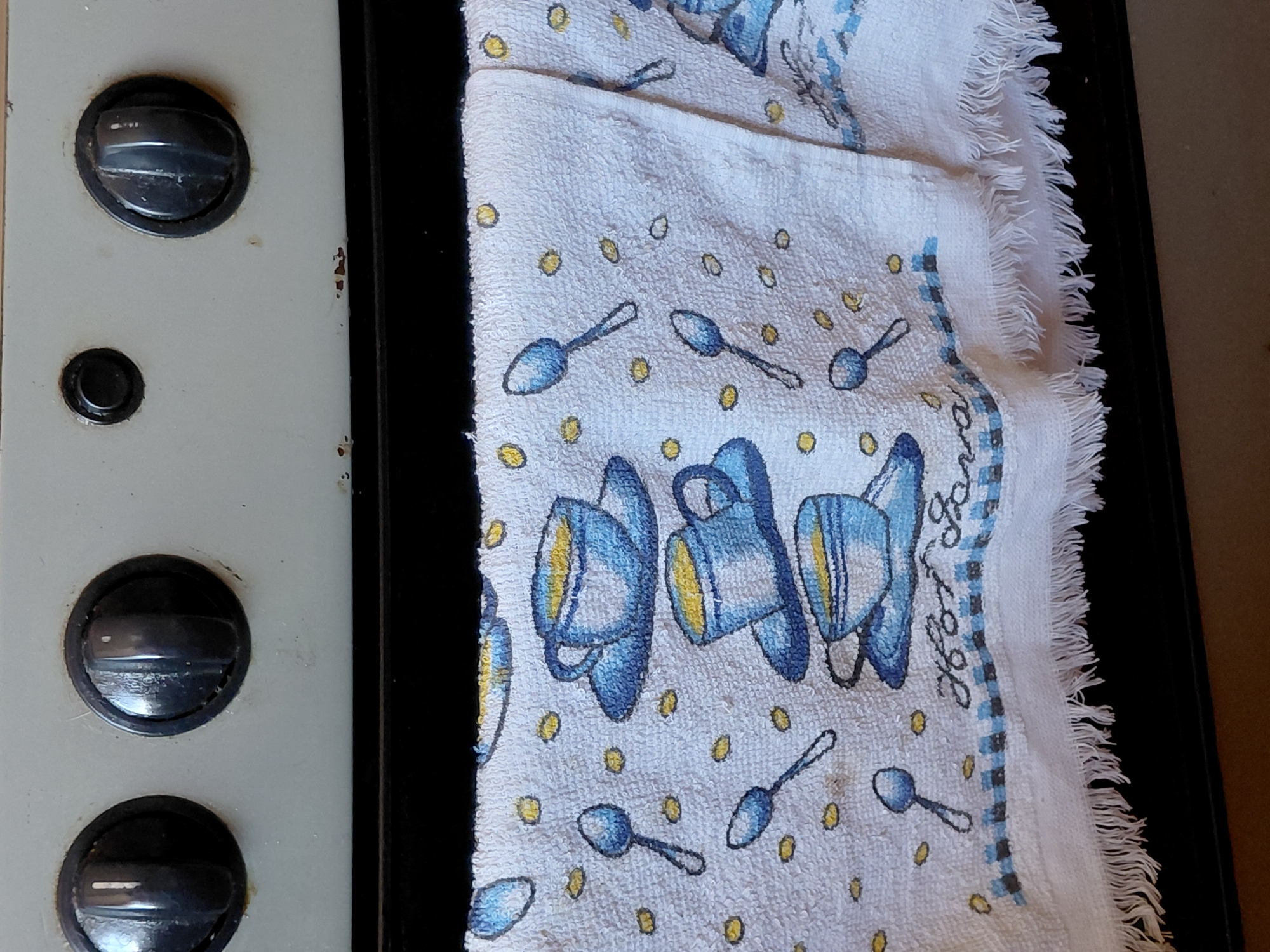 The image shows a close-up view of a white kitchen towel hanging over the edge of a stovetop. The towel is decorated with a pattern of blue and yellow coffee cups and spoons, along with scattered yellow dots that appear to be coffee beans. The bottom edge of the towel has a fringe. The stovetop itself is light gray and has four black knobs visible. The towel is draped over the front of the stove, obscuring most of the appliance. The background is dark and out of focus. There are no people or any indication of the location being Santa Ana, Venezuela.