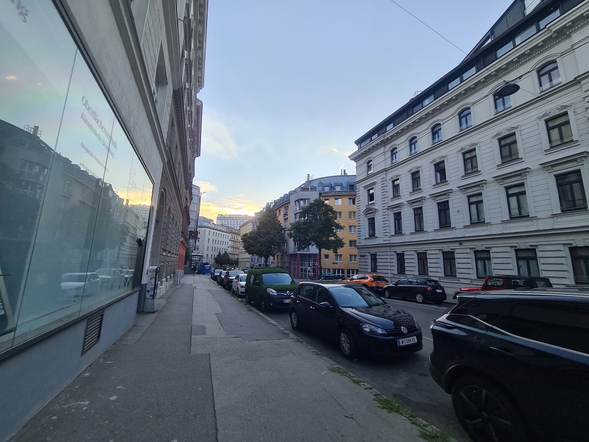 The image depicts a street scene in Vienna, Austria, at what appears to be late afternoon or early evening. The perspective is from a low angle, looking down a street lined with buildings on both sides. On the left, a modern building with a large glass facade reflects the street and the sky. Text in German is visible on the glass, including "Oberflächentechnik" and "Künstlerische Gestaltung".
On the right side of the street, a row of older European-style buildings with multiple stories and many windows are visible. The buildings have classical architectural details.
The street itself is paved, with a sidewalk running alongside the buildings on the left. Several cars are parked along the street, including a dark colored Volkswagen Golf with a license plate starting with "W", a dark colored compact van with a green, grass-like covering, and other vehicles of various makes and models. Some trees are planted along the sidewalk.
In the distance, the street recedes, and more buildings and cars are visible. The sky is clear with some light clouds, and the setting sun casts a warm glow, particularly visible in the reflections on the glass facade and towards the horizon. There are no people visible in the image. The scene is static, with parked cars and buildings.
