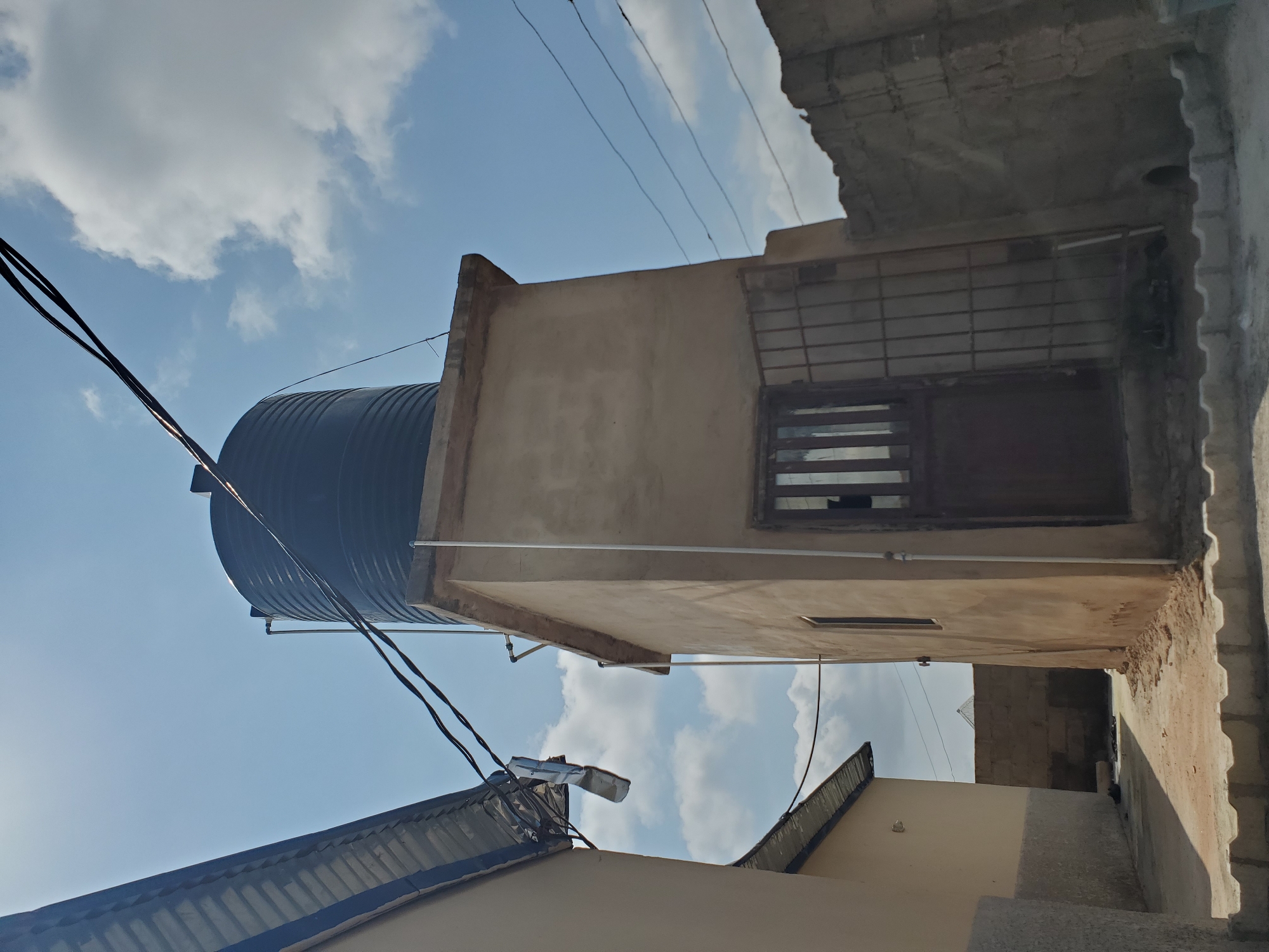 The image depicts an outdoor scene in the city of Kason, Nigeria. In the foreground, a paved walkway with decorative concrete edging leads towards a tan-colored structure. This structure has a door with a metal grate and a small window. Above this building, a large black water tank is visible. Overhead, power lines crisscross the sky, which is partly cloudy. To the left, the corrugated metal roof of another building is visible, leaning towards the right. To the right of the tan structure, a rough stone wall forms a boundary. The scene is illuminated by natural daylight. There are no people visible in the image.