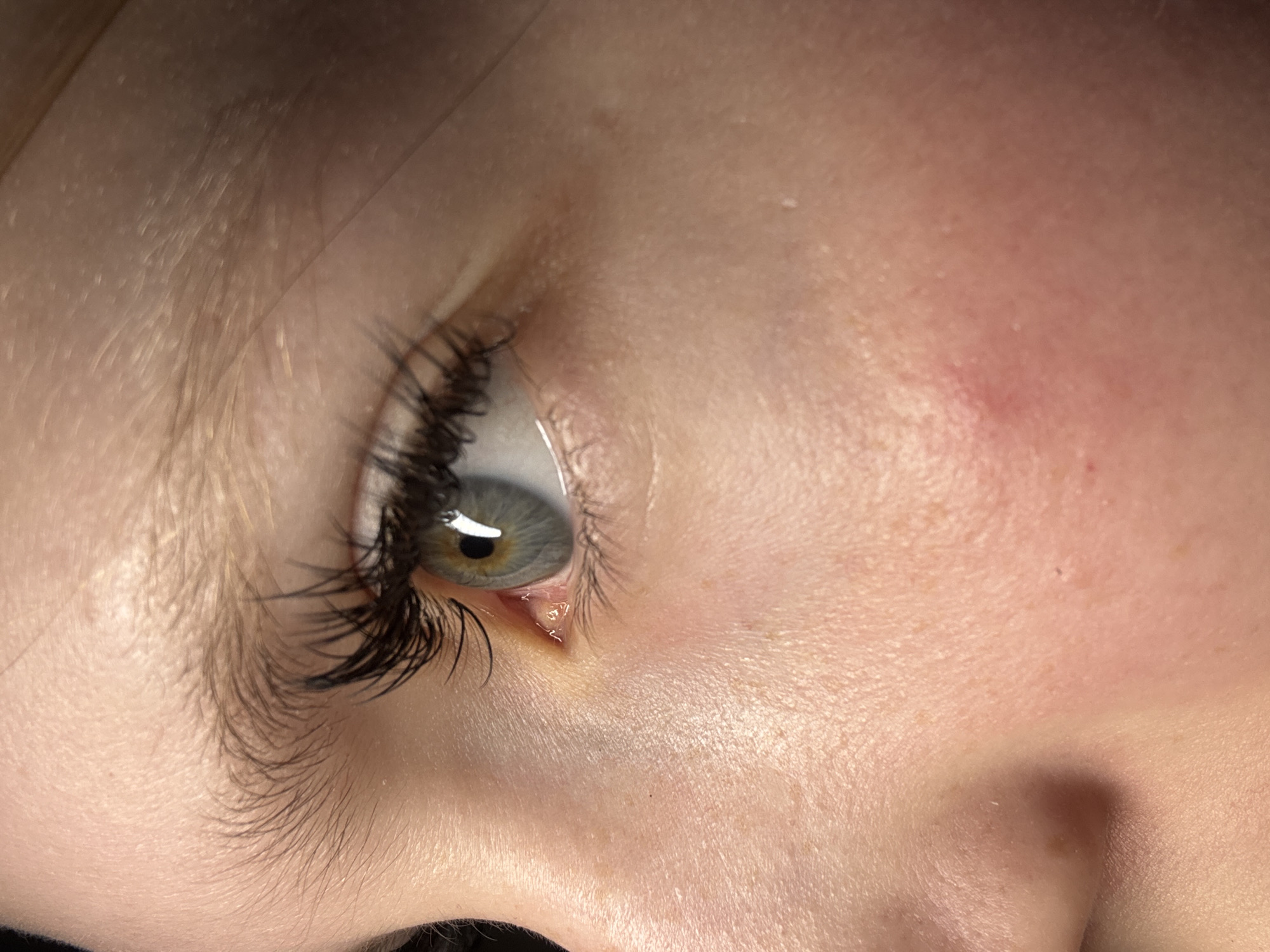 A close-up image depicts a human eye, exhibiting long, dark eyelashes and a light green iris. The surrounding skin is fair with visible pores and a small red mark on the cheek. There is no evidence to suggest this image was taken in Chaikovsky, Russia, or anywhere else specifically.