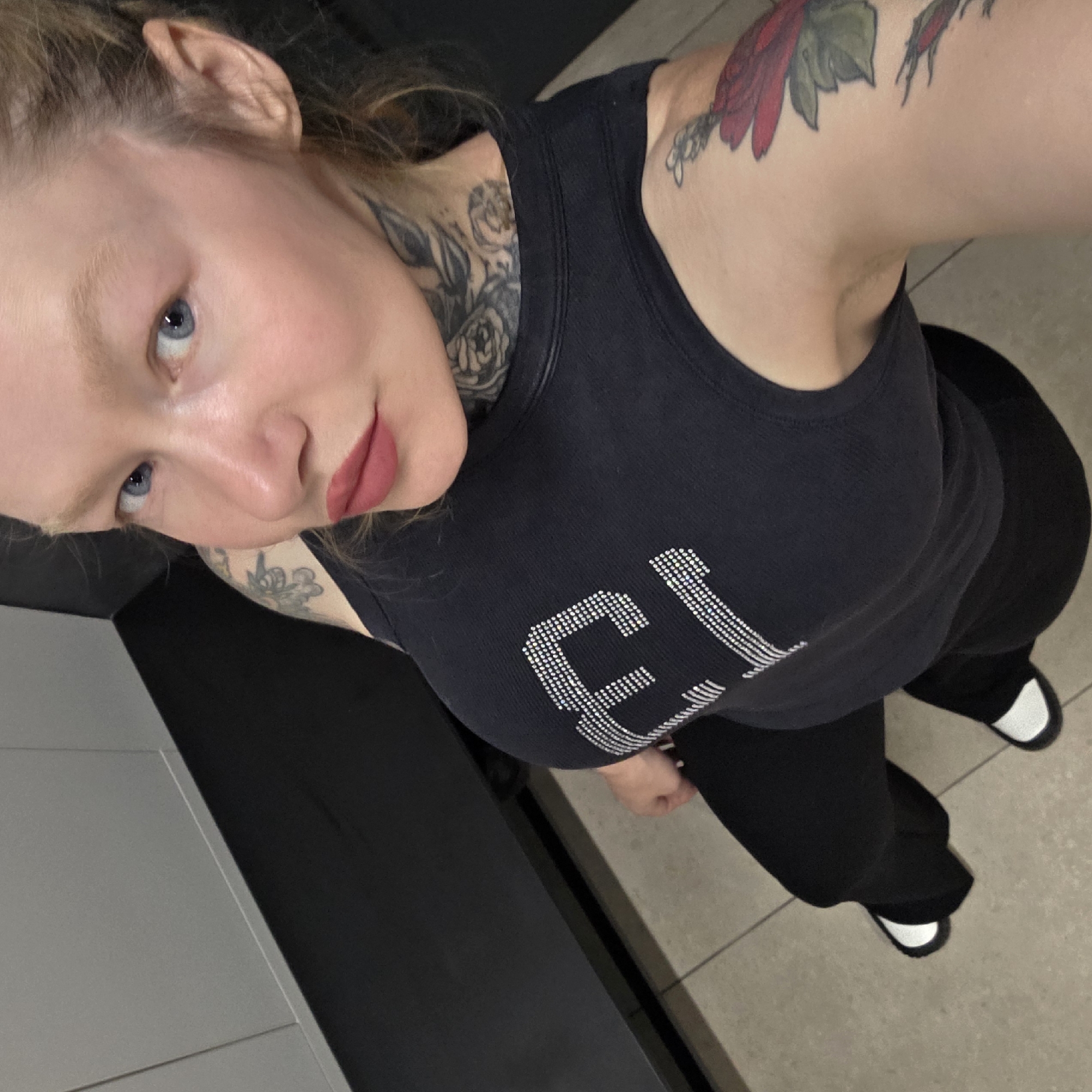 A fair-skinned woman with blue eyes and shoulder tattoos is pictured in a selfie taken from above. She is wearing a black tank top with rhinestones on the front, black pants, and black shoes with white soles. The woman has a neck tattoo. The background suggests she is indoors, possibly in a locker room or similar space, with beige tiles and a grey wall or locker to the left. The exact location within Saint Petersburg, Russia, is not discernible from the image.