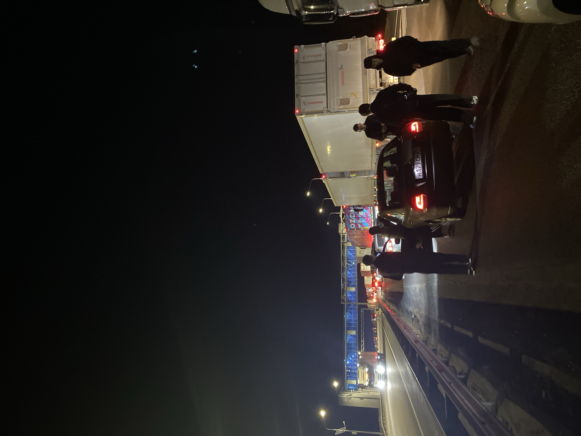 At night in Iksuar, Russia, several individuals are gathered around a dark-colored car with a license plate reading "H 179 CM 777" positioned in a line of vehicles on a roadway. The cars and trucks are stopped at what appears to be a checkpoint with illuminated blue gantries overhead. Some of the individuals are wearing dark clothing, and some have face coverings. A large white truck with the word "SUNHUNK" printed on it is visible adjacent to the car. Streetlights are interspersed along the roadway. The overall scene suggests a border crossing or security inspection point.
