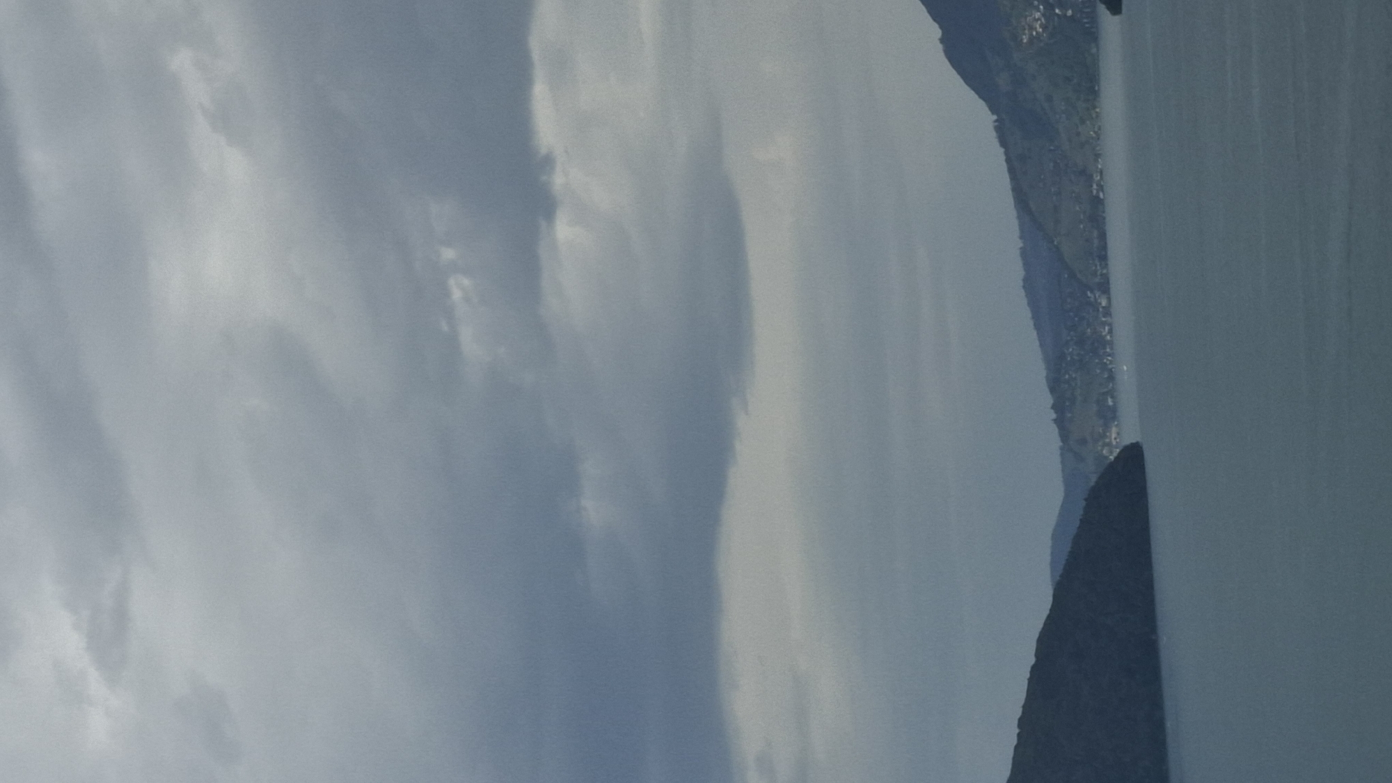 The image shows a landscape view of Lake Lucerne with mountains and a town on the shore. The sky is overcast with heavy clouds. In the foreground, a calm, blue body of water dominates the lower half of the frame. To the left, a dark, tree-covered mountain slopes down to the water. To the right, across the lake, a densely populated hillside town is visible, nestled against a green mountain. Buildings and roads are discernible, and a few boats are scattered on the water. The overall atmosphere is subdued and somewhat gloomy due to the cloudy weather. The image appears to be taken from an elevated viewpoint, possibly a hillside or a boat, looking across the lake towards Beckenried, Switzerland.