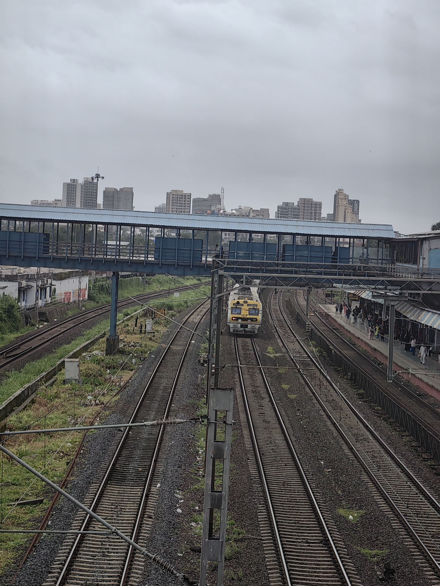 The image shows a busy railway station in Mumbai, India, under an overcast sky. A yellow train is approaching the platform on the left side of the tracks. A pedestrian overpass, painted blue, spans across the railway tracks, connecting different parts of the station. Several people are visible on the platform and within the overpass. Buildings and skyscrapers form the city skyline in the background, suggesting an urban environment. The railway tracks are well-maintained, with ballast filling the spaces between the sleepers. Overhead electrical wires are suspended above the tracks, powering the electric train. The scene depicts a typical day at a bustling Indian railway station.