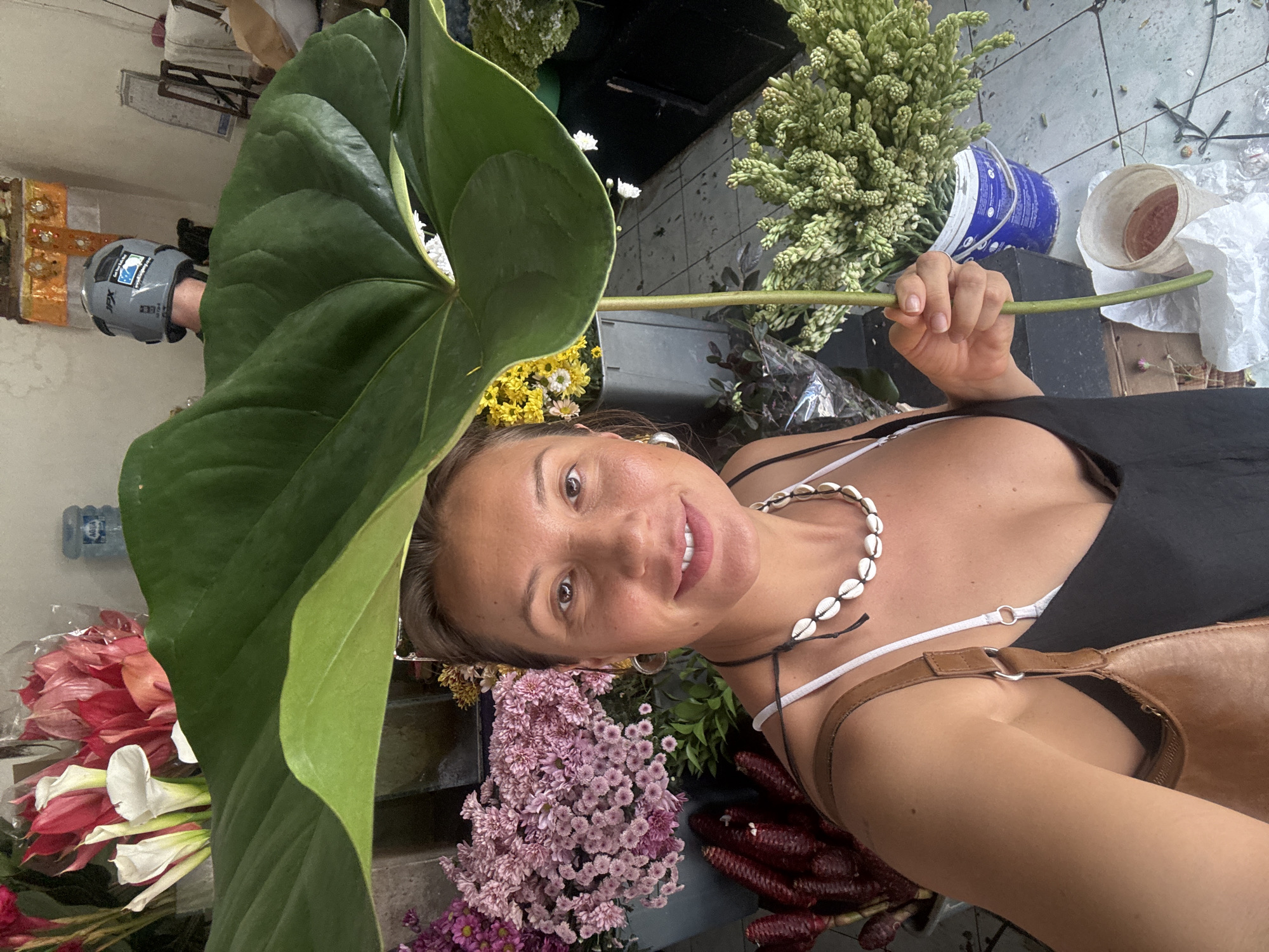 A selfie captures a woman, seemingly within a floral setting. She is positioned among a variety of flowers, including pink chrysanthemums, red dahlias, and white calla lilies. She holds a large, green leaf in front of her face. She is wearing a black tank top, a white bikini top, and a necklace made of seashells. A tan leather bag is positioned over her right shoulder. In the background are green flowers, a blue bucket and other objects. The location is identified as being in North Kuta, Indonesia.