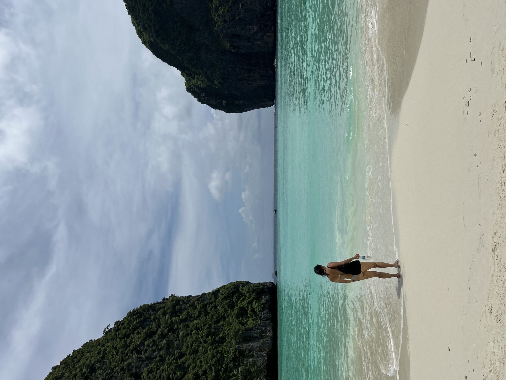 A lone woman stands on a white sand beach in Ao Nang, Thailand, facing the turquoise water. She is wearing a dark swimsuit and carrying a black bag over her shoulder, with a water bottle in her right hand. The beach is calm with gentle waves lapping at the shore. On either side of the bay, large, verdant limestone cliffs rise dramatically from the water, covered in lush green vegetation. The sky above is partly cloudy with a mix of grey and white clouds, and the water is clear and vibrant. In the distance, several small boats are visible on the horizon.